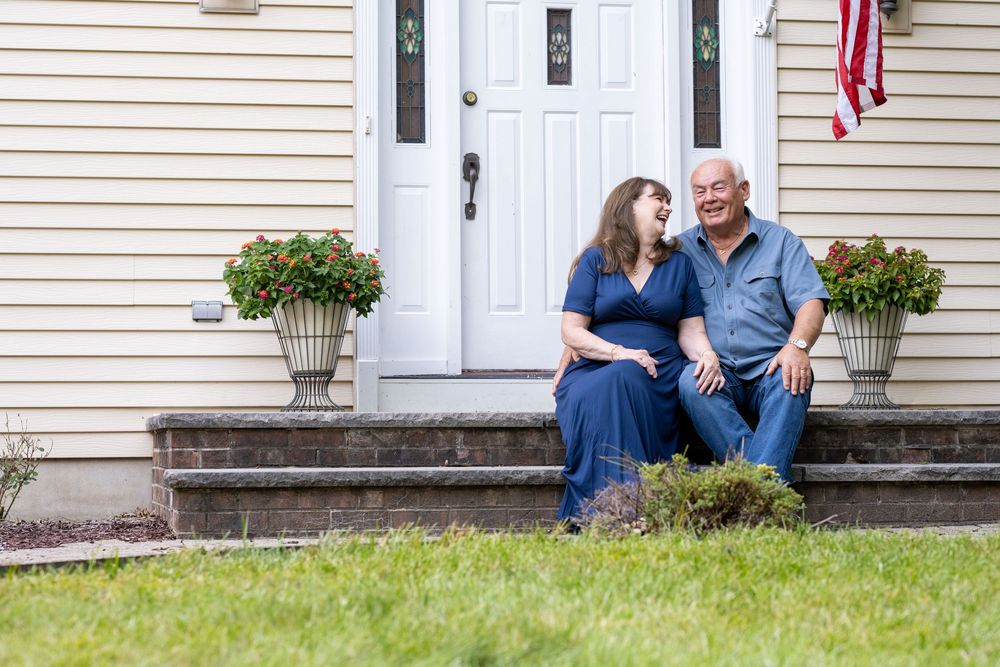 A couple sitting together on front porch steps with potted flowers and an American flag.