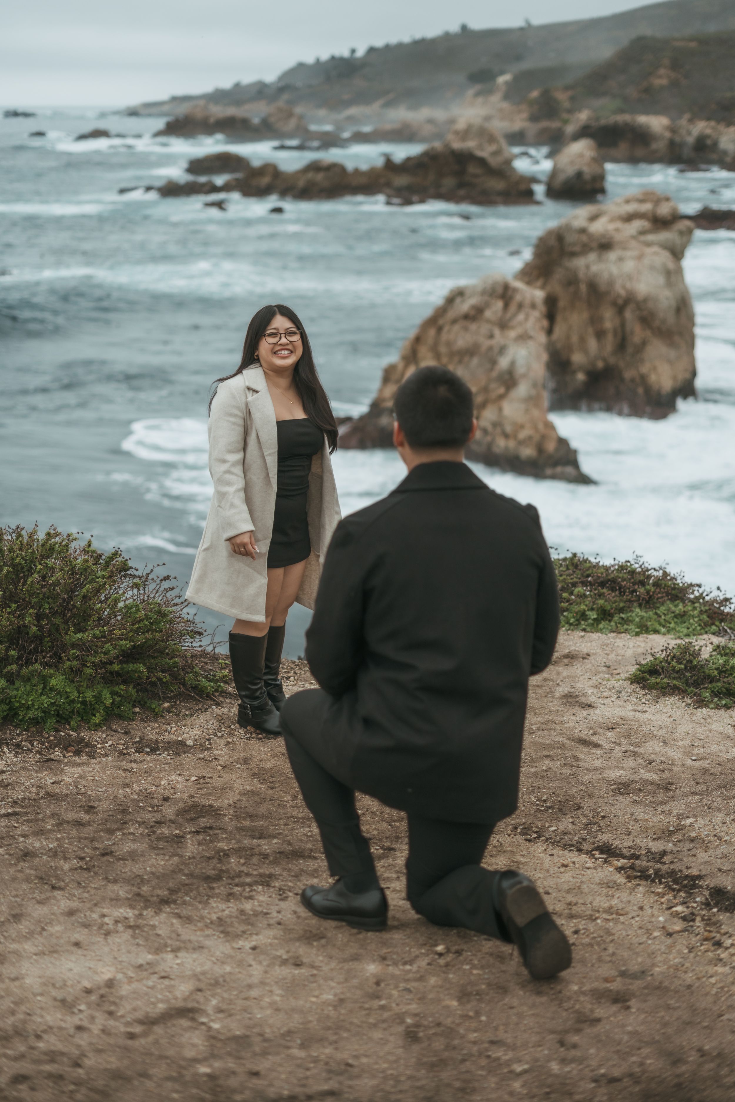 A couple shares a romantic proposal moment on a cliff overlooking the rocky Pacific coast on a cloudy day.