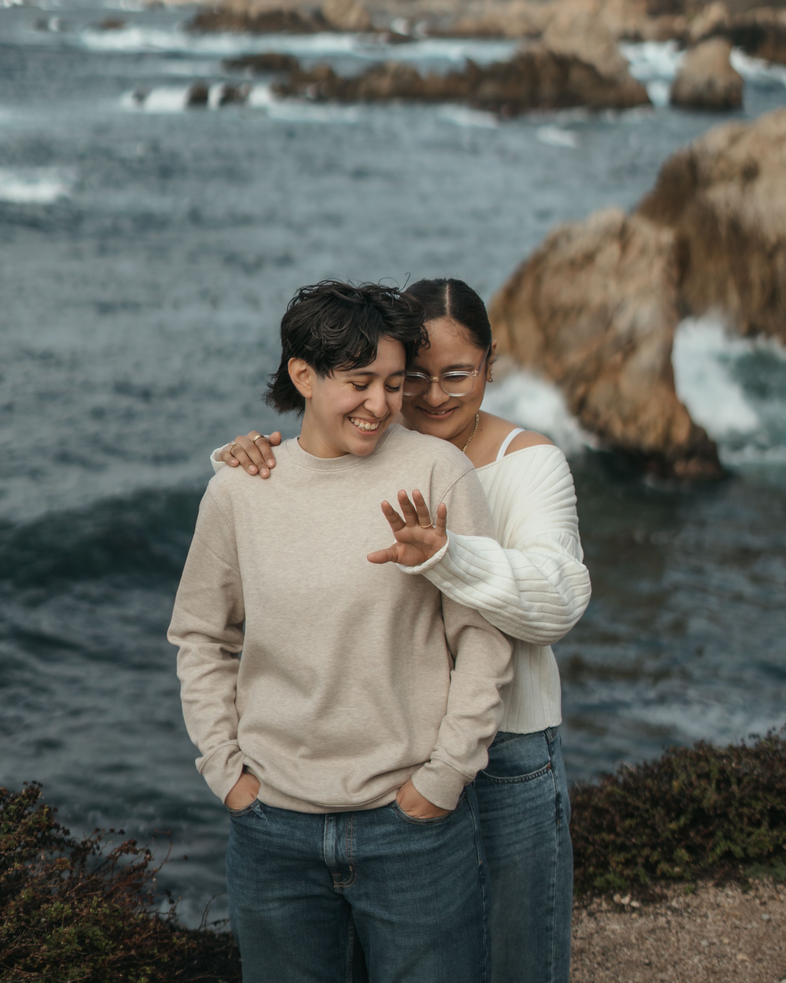 Two people share a tender embrace while standing on rocky coastal cliffs with the ocean waves crashing behind them.