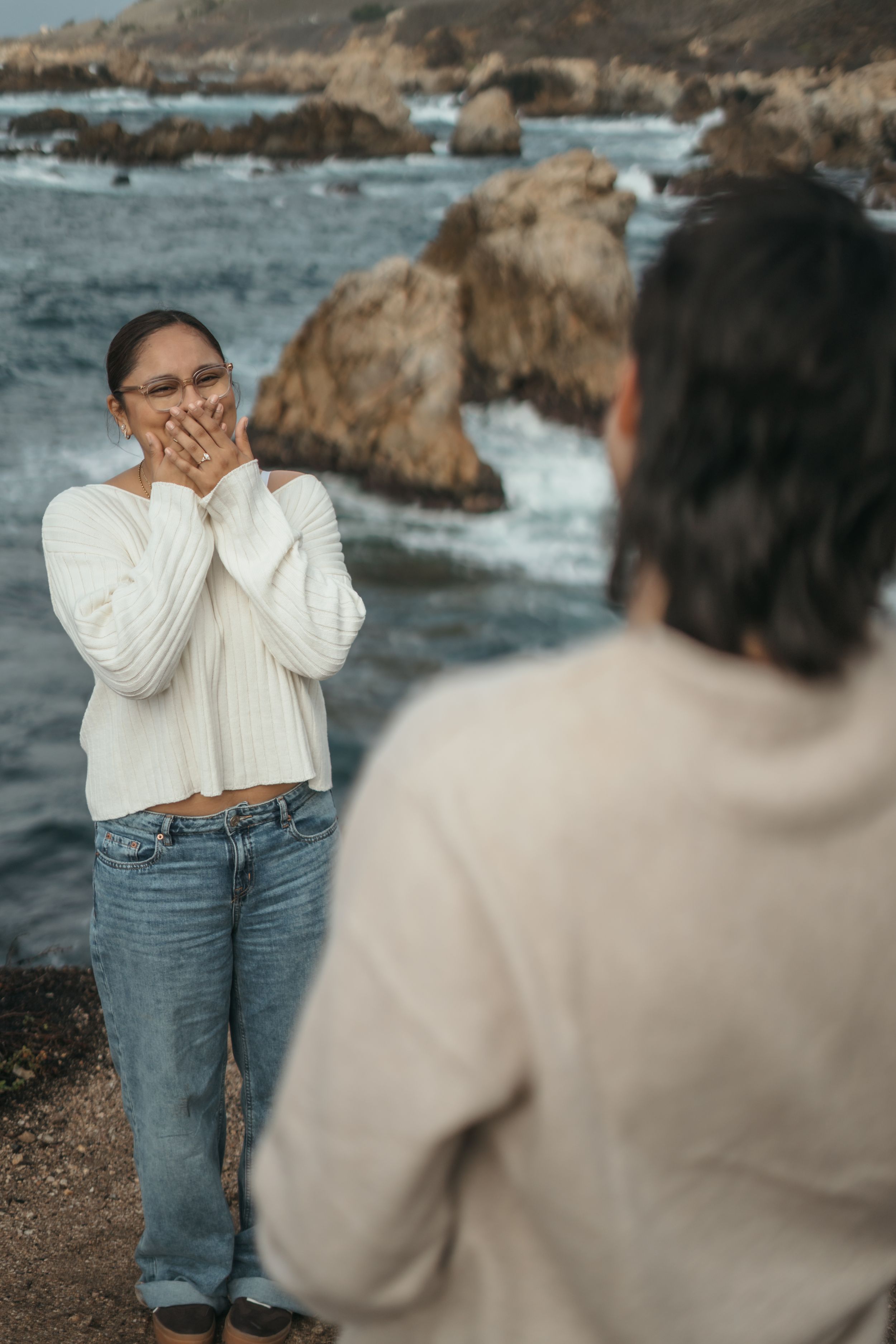 Couple sharing a romantic moment on rocky coastal cliffs during sunset with ocean waves crashing in background.