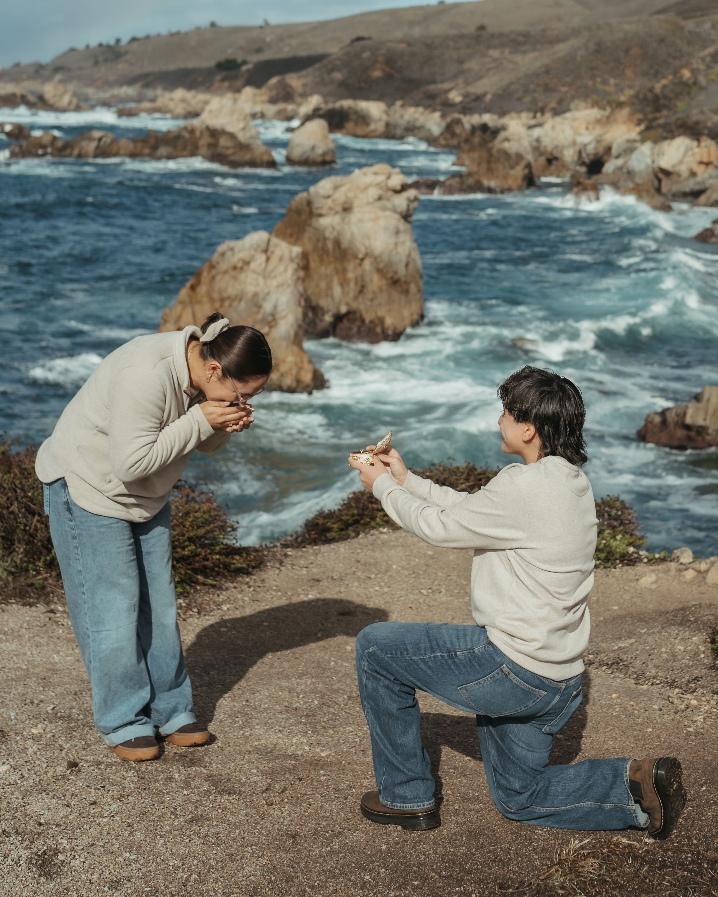 A couple shares a tender moment on rocky coastal cliffs overlooking crashing ocean waves in Big Sur.