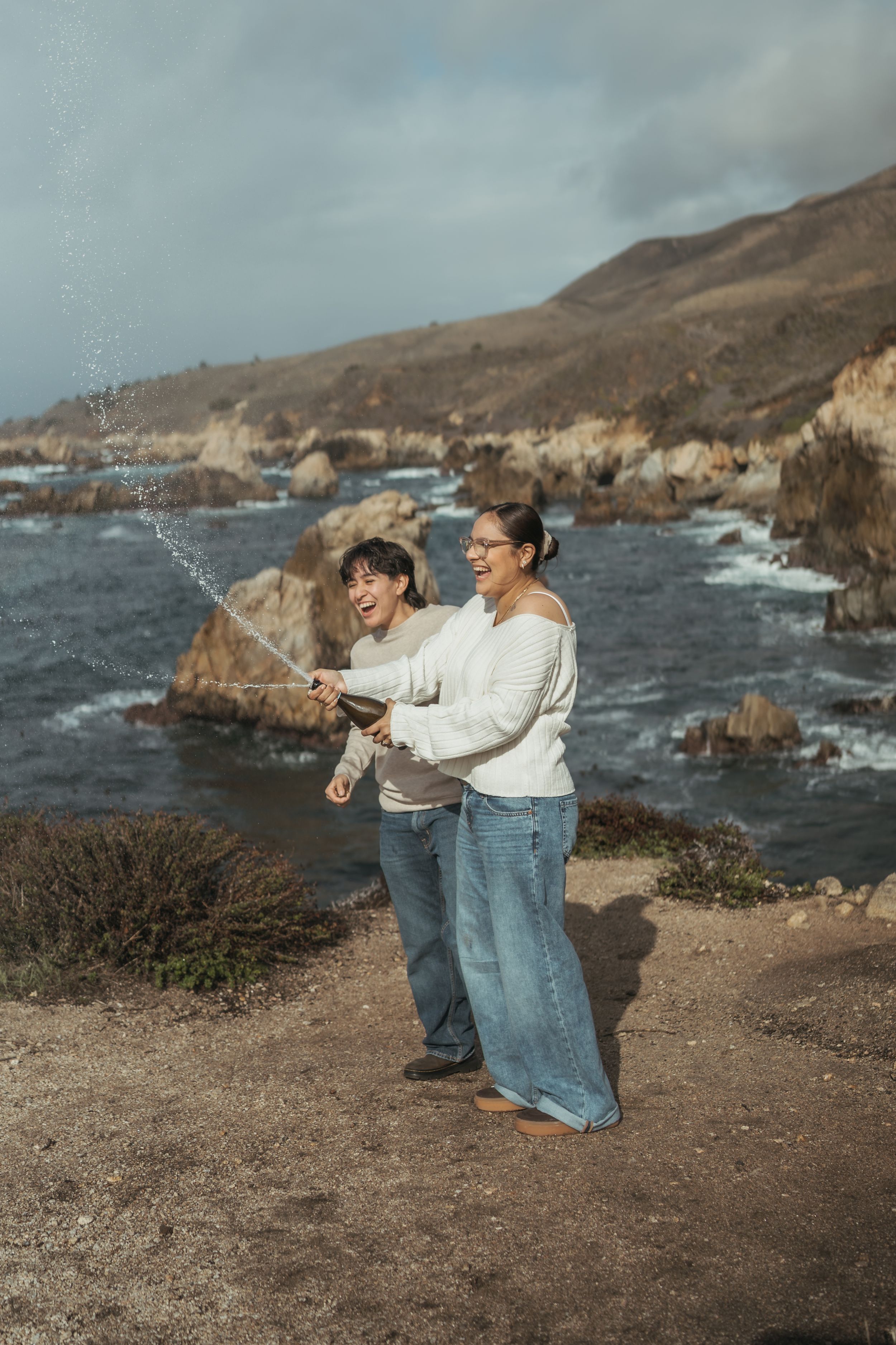 A couple stands together on a coastal cliff overlooking the rocky Pacific Ocean shoreline at sunset.