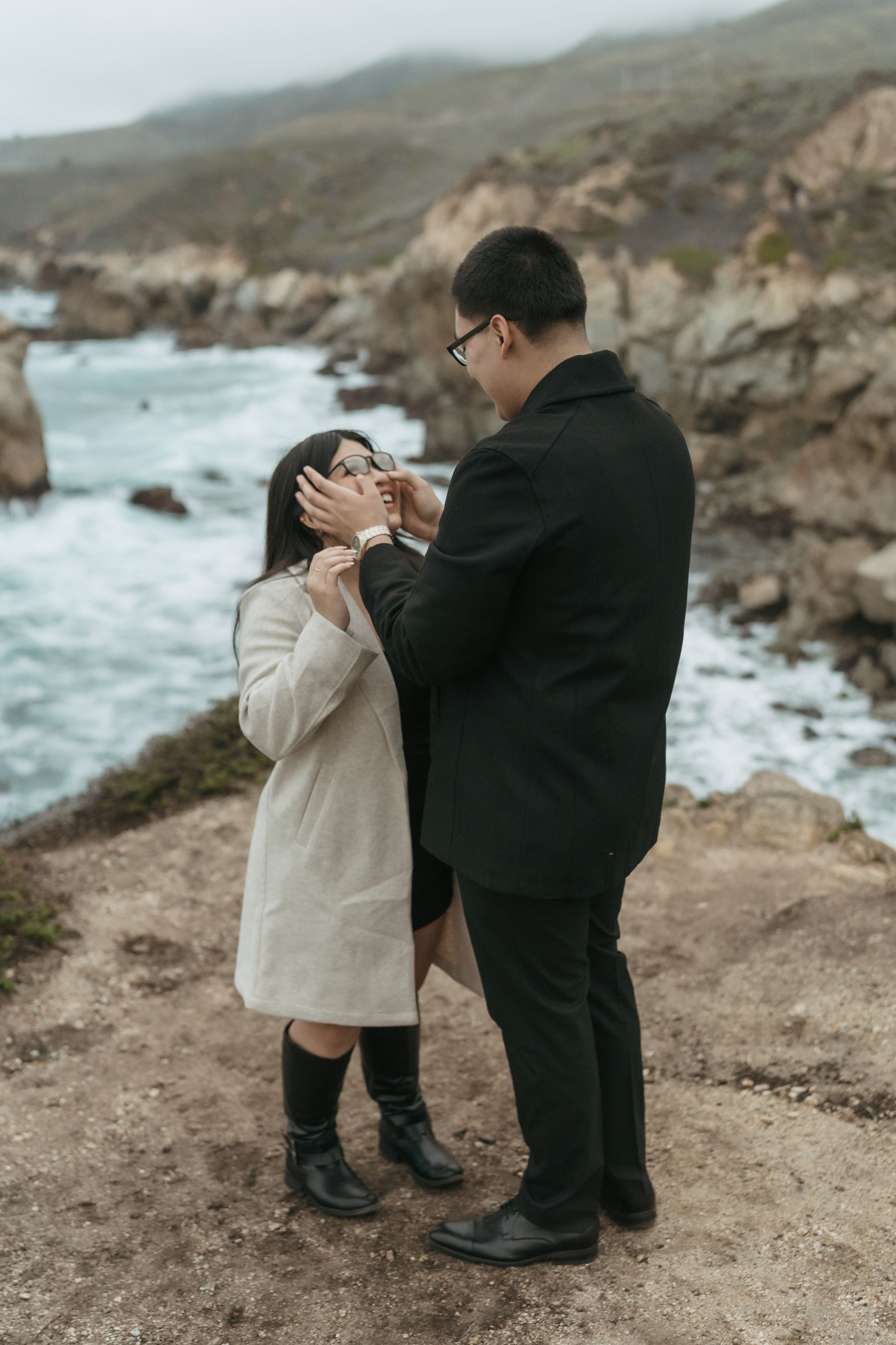 A couple shares a tender moment on a rocky coastal cliff overlooking dramatic ocean waves and rugged shoreline.