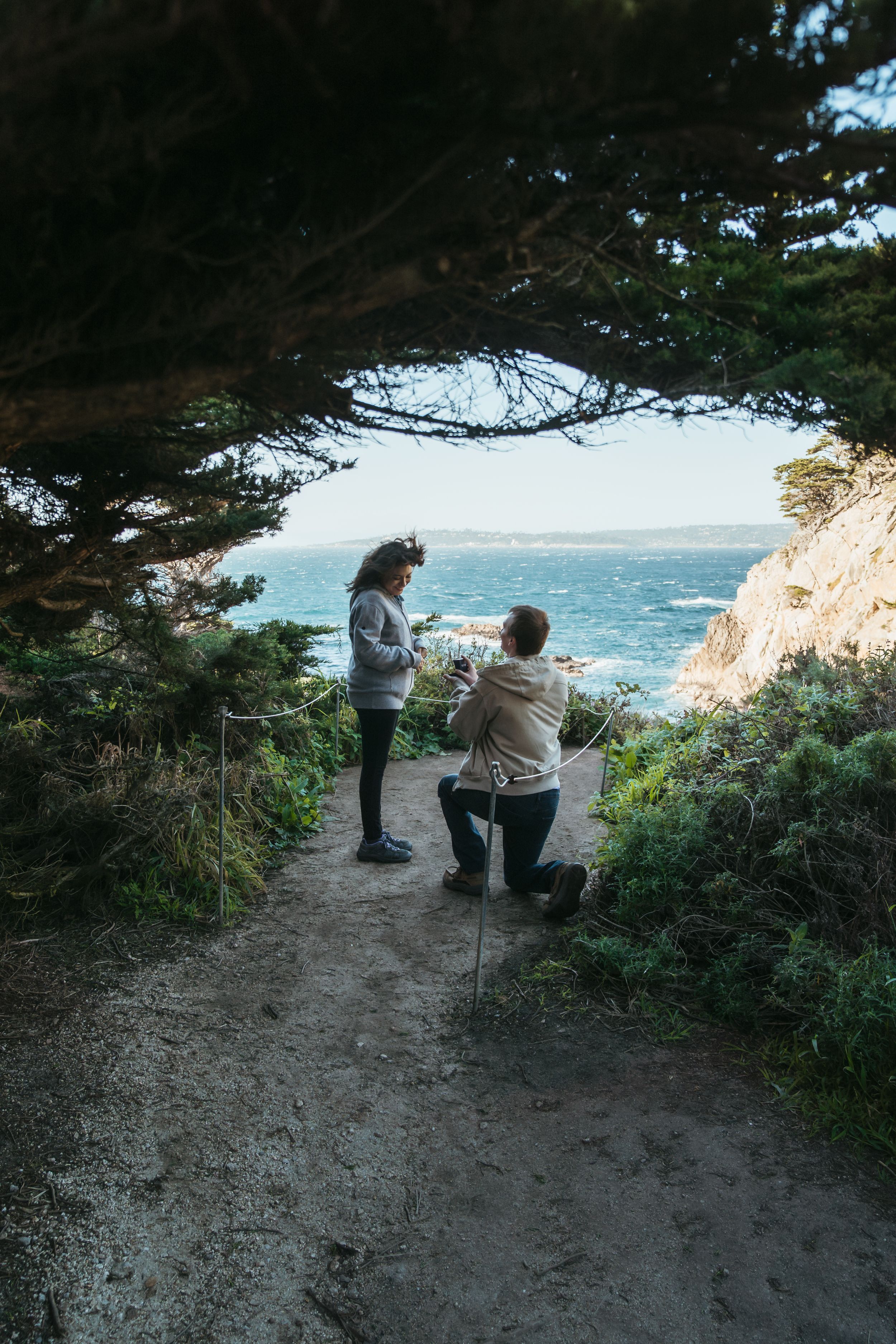Romantic proposal scene unfolds on a scenic coastal cliff overlooking the ocean on a sunny afternoon.