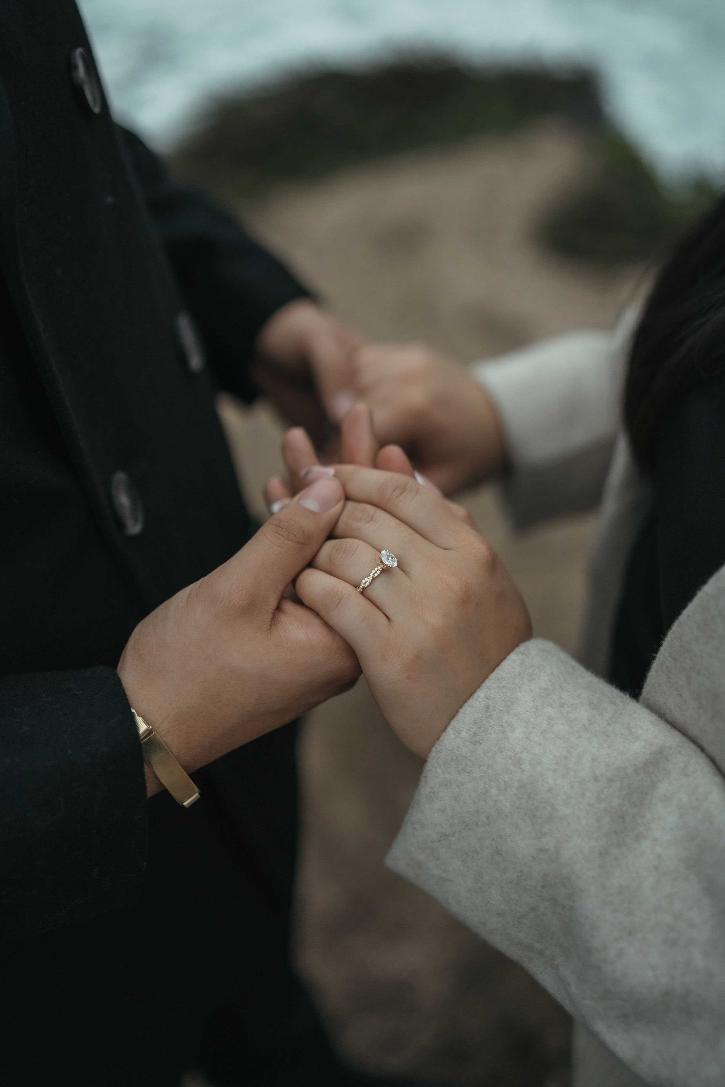 Close-up of hands holding together with engagement ring showing, wearing black and gray clothing in moody lighting.