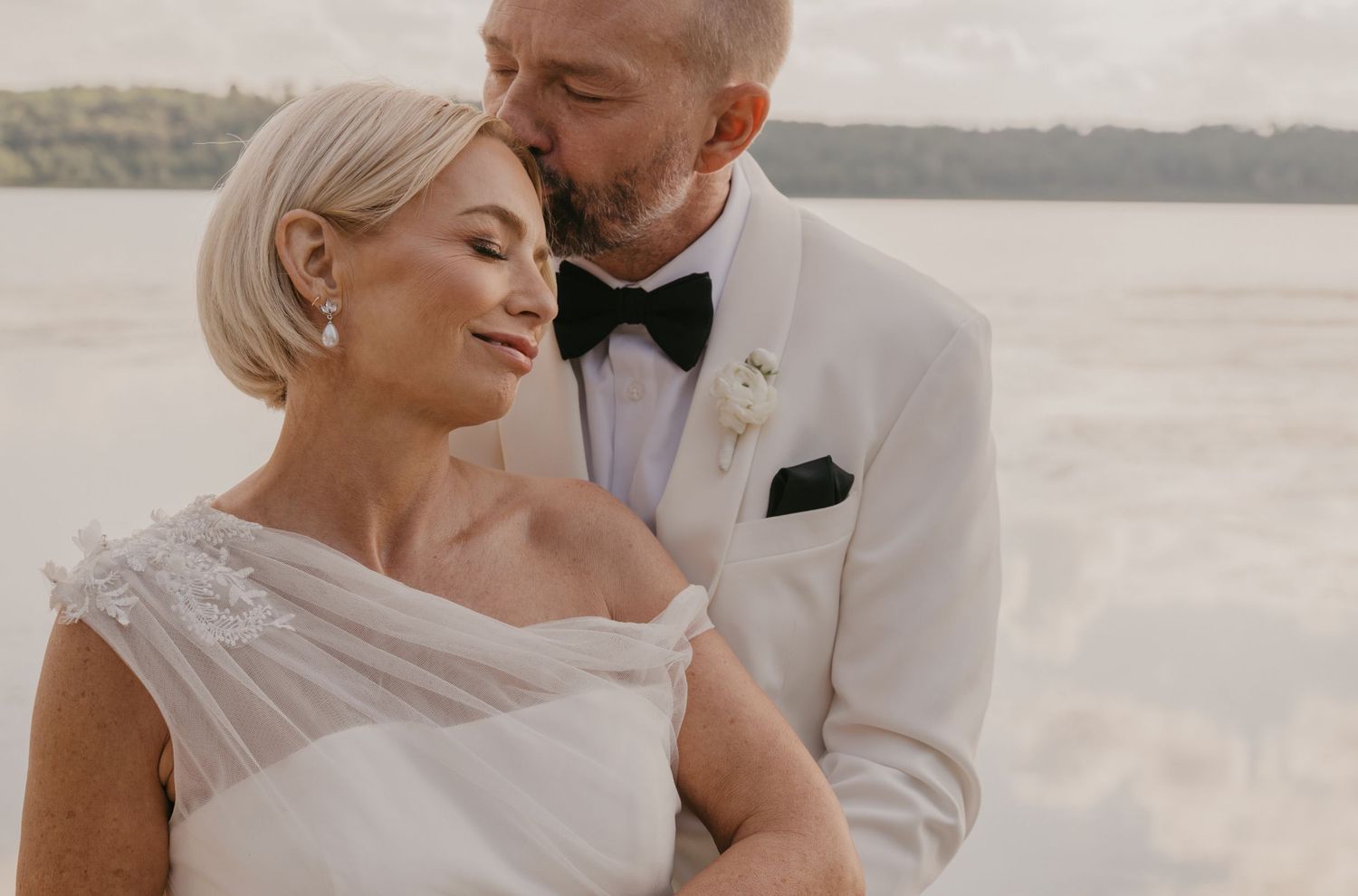 Elegant couple in wedding attire share an intimate moment by a serene waterfront at sunset.