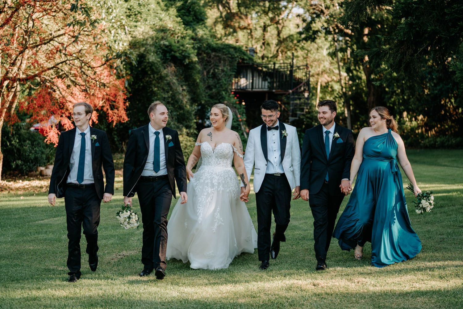 Wedding party walks together on grass path through autumn trees with golden sunlight filtering through branches.