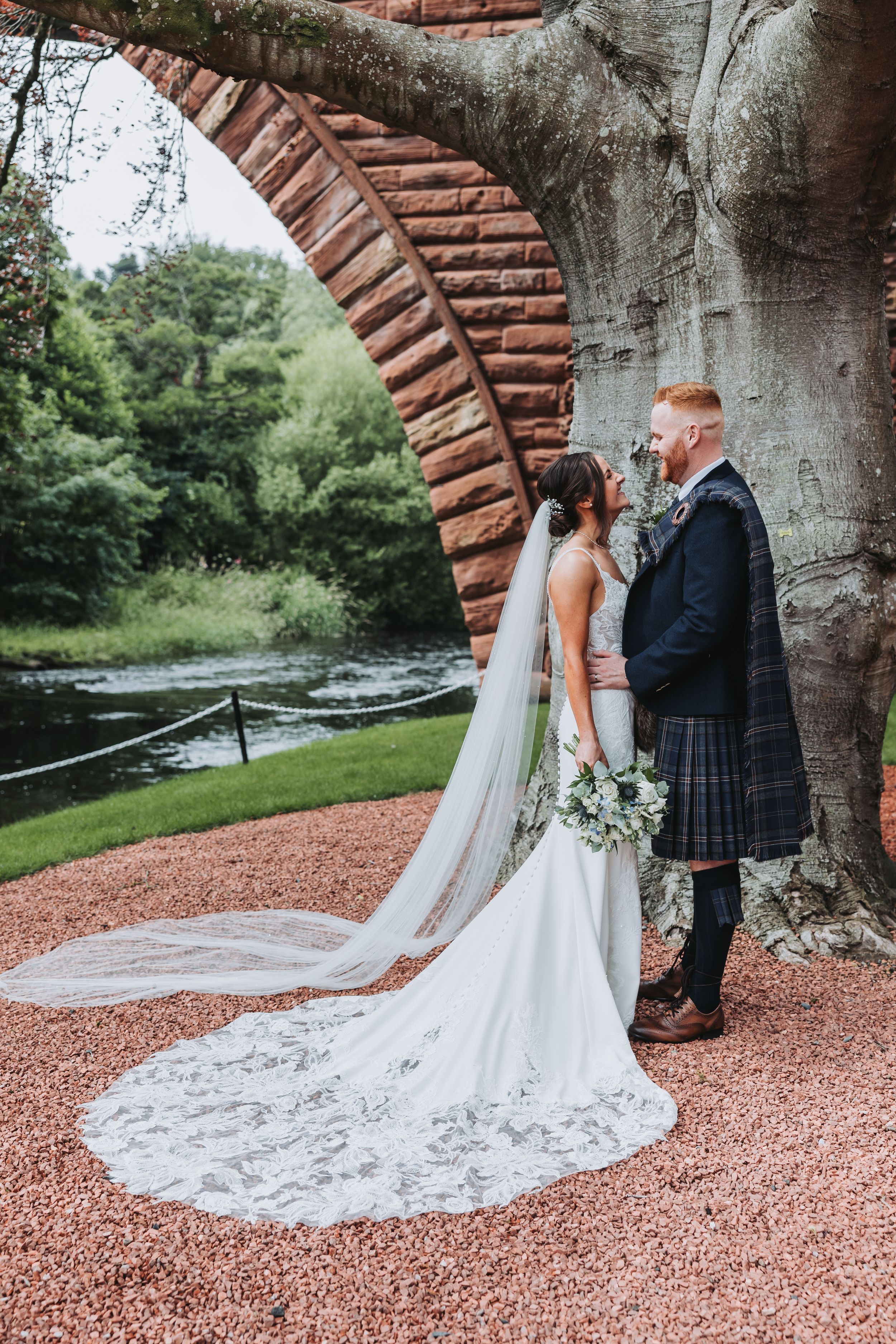 Wedding couple poses under rustic brick archway on red gravel path.