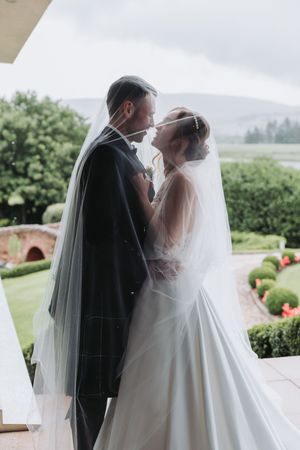 A romantic wedding couple shares an intimate moment on a porch with lush greenery in the background.