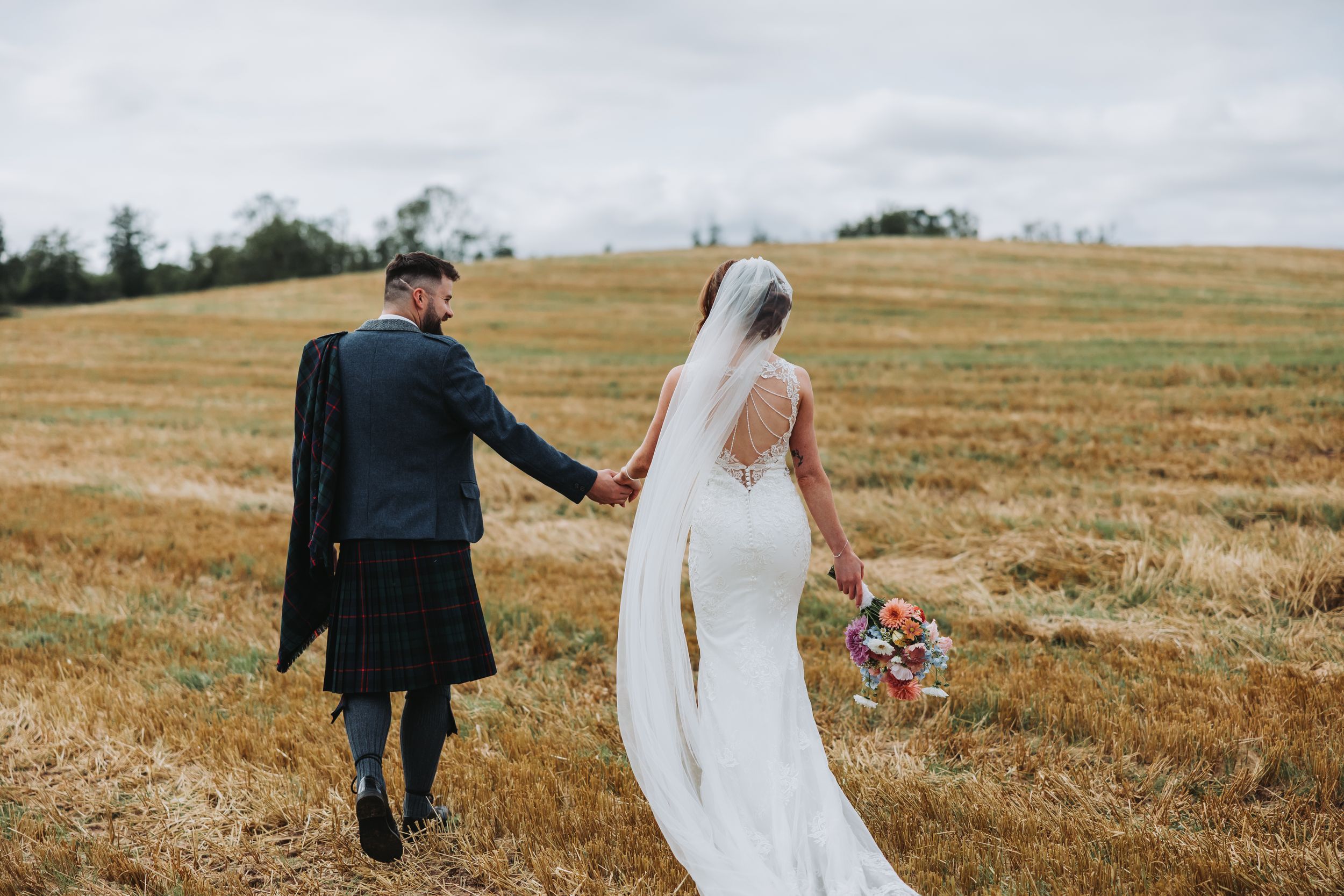Newlyweds walking through golden wheat fields under cloudy skies.