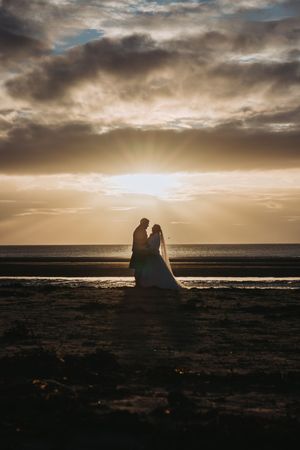 Silhouetted wedding couple against a dramatic sunset sky on a beach.