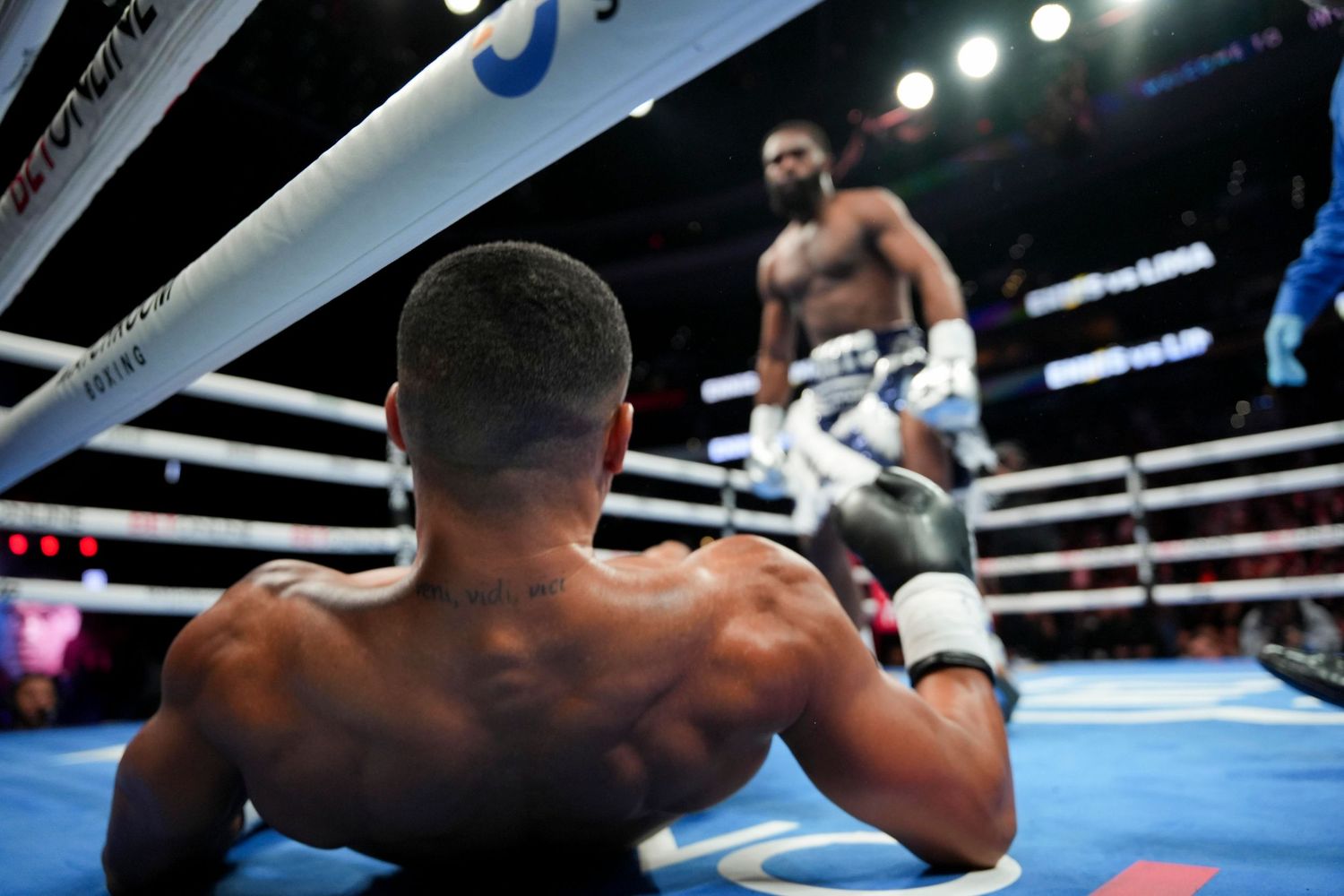 A knocked-down boxer kneels on the canvas while his opponent stands ready in a professional match.