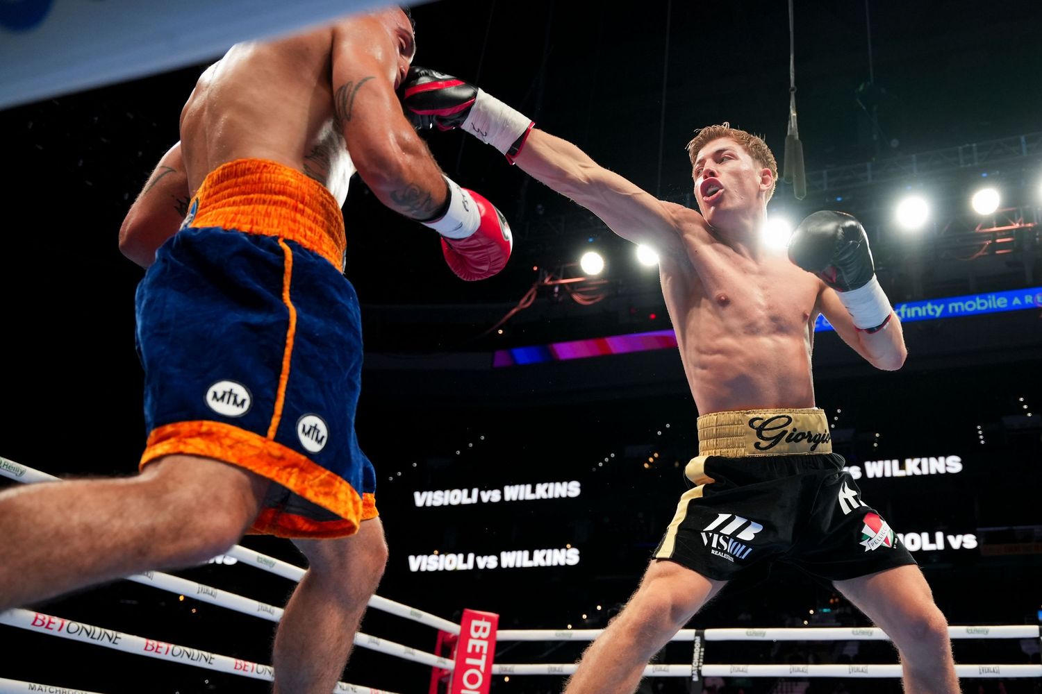 Two boxers exchange punches in a professional boxing match, with ring banners visible in the background.