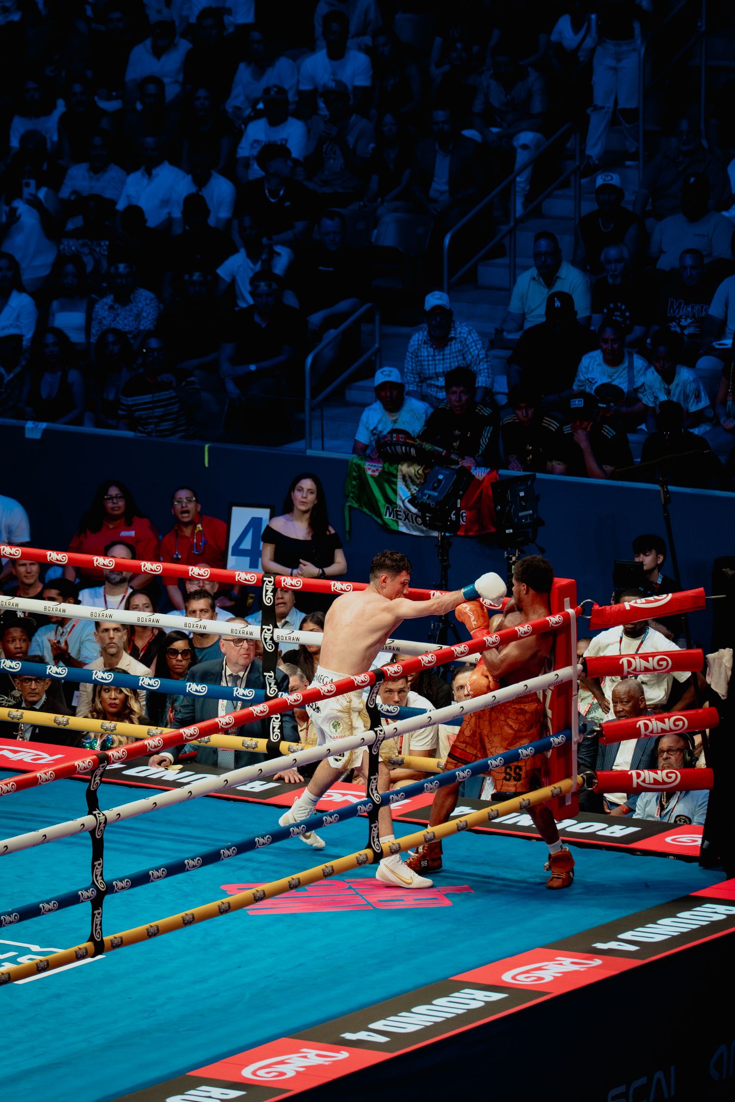 Two boxers exchange punches near the ropes in a packed professional boxing arena with colorful lighting.