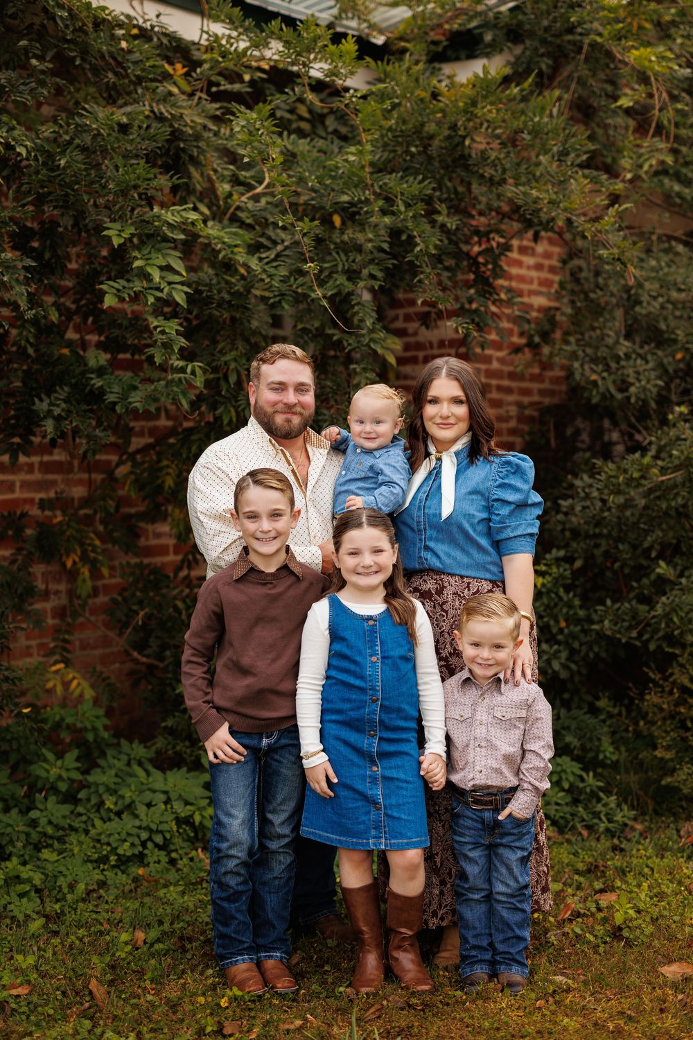 Family poses together in coordinated casual outfits against brick and foliage backdrop.