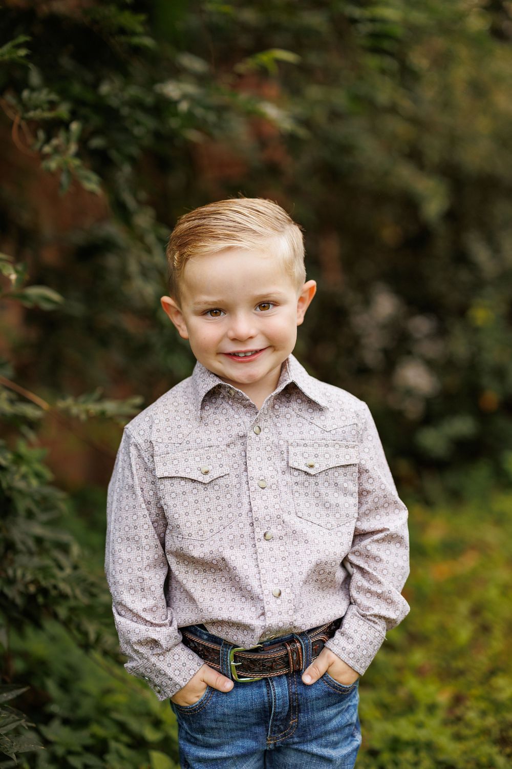 Child in patterned button-down shirt and jeans smiles while standing in garden setting.