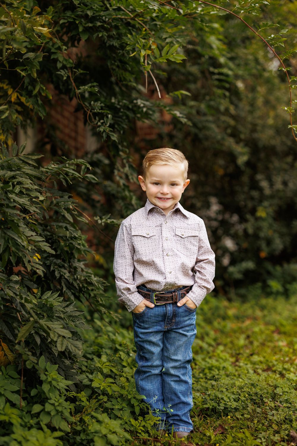 Young child in light button-up shirt and jeans poses among green foliage.