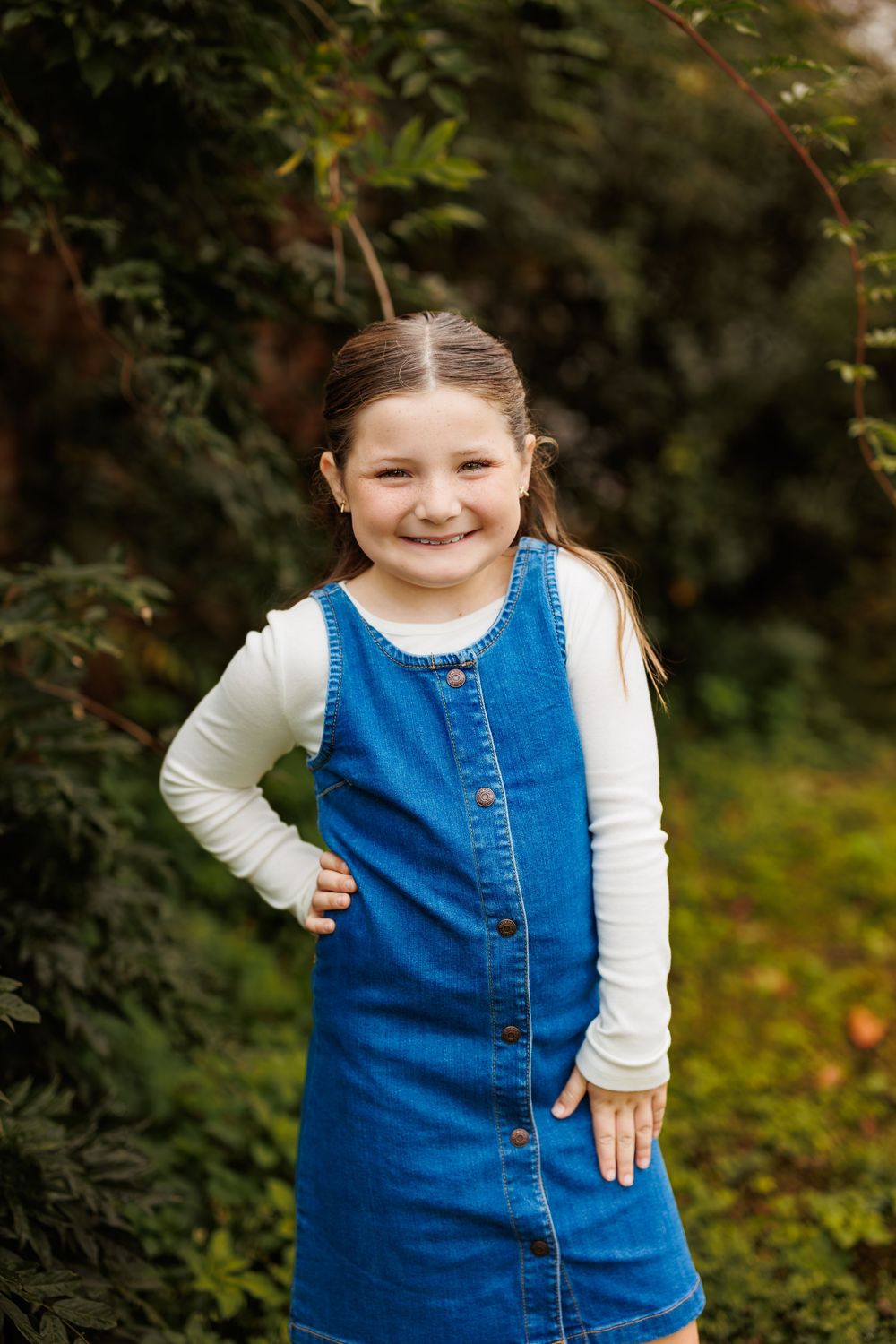 Child in denim jumper dress and white sweater smiles in outdoor garden setting.