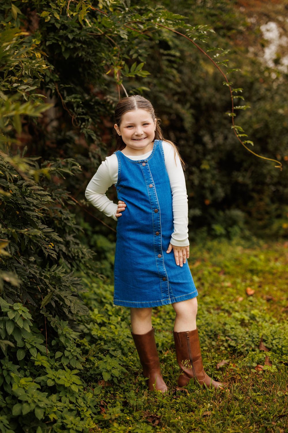 Child in denim jumper dress and white shirt plays in garden setting.