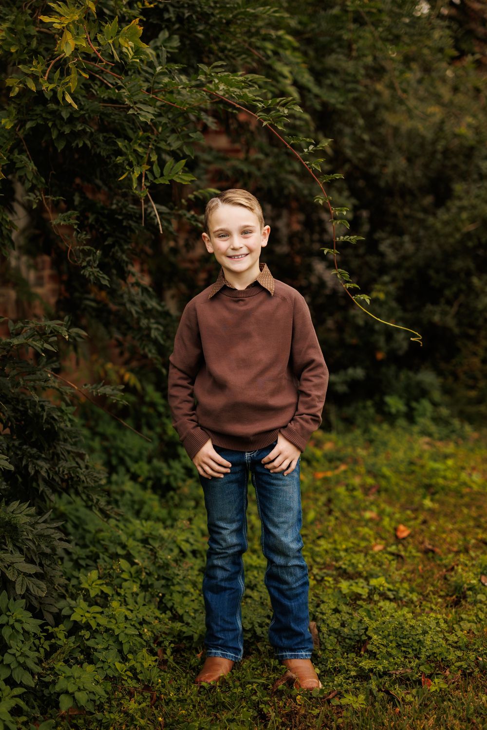 Child in brown sweater and jeans poses casually in autumn garden setting.