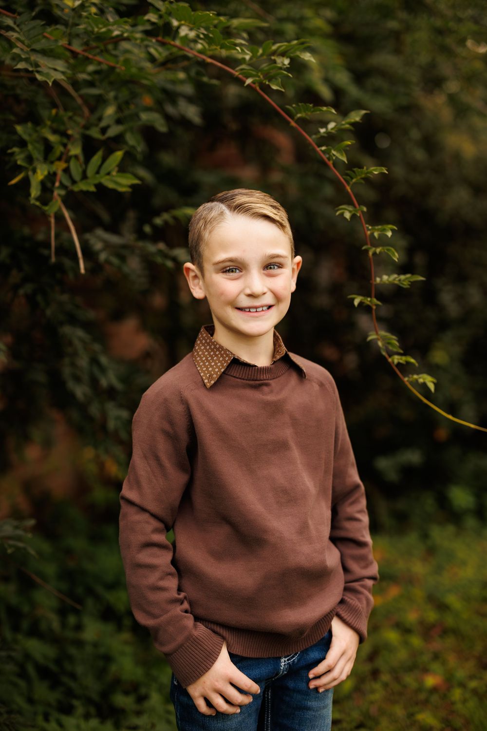 Young child wearing brown sweater poses outdoors against natural foliage background.