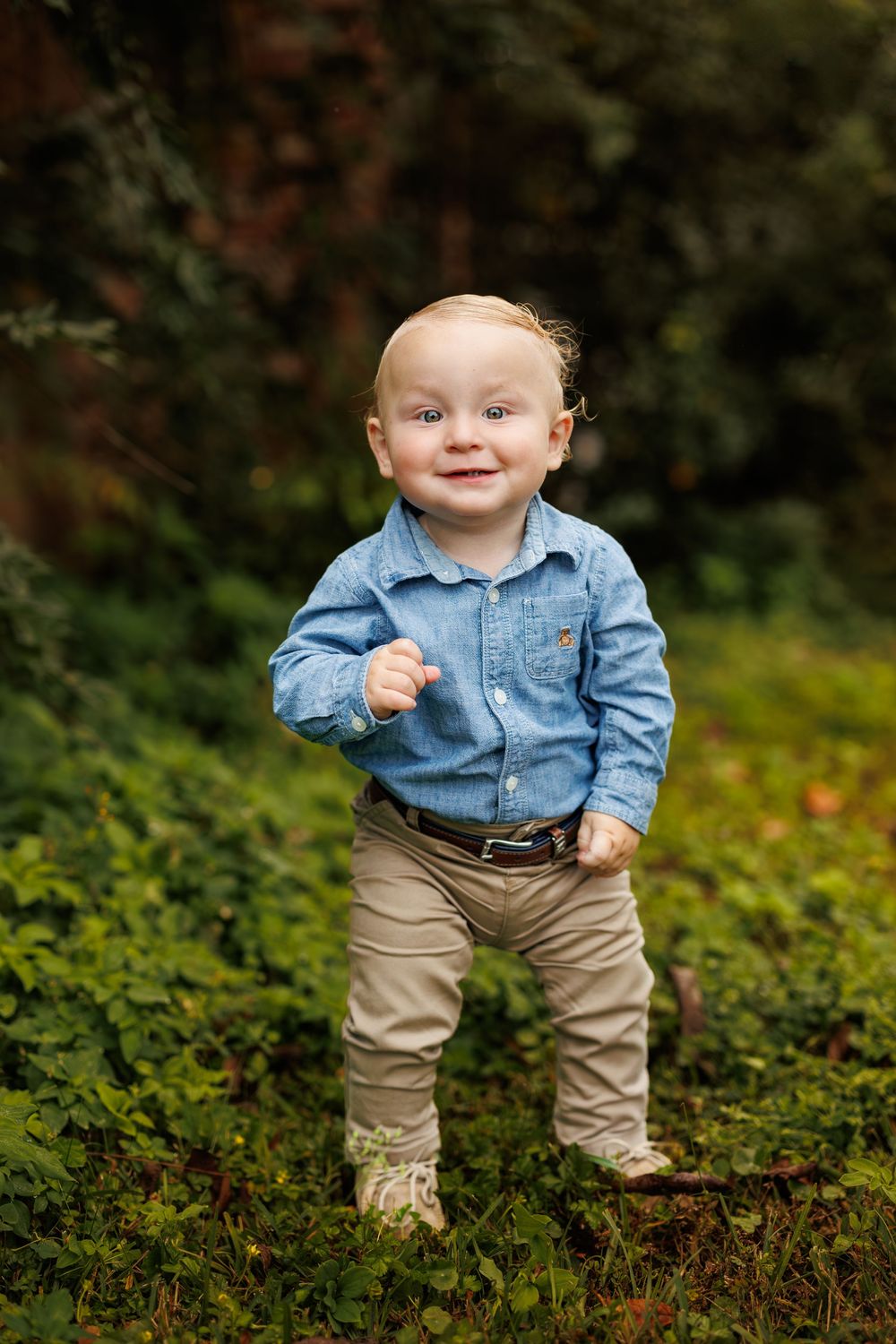 Small child in denim shirt and khaki pants stands on mossy ground outdoors.