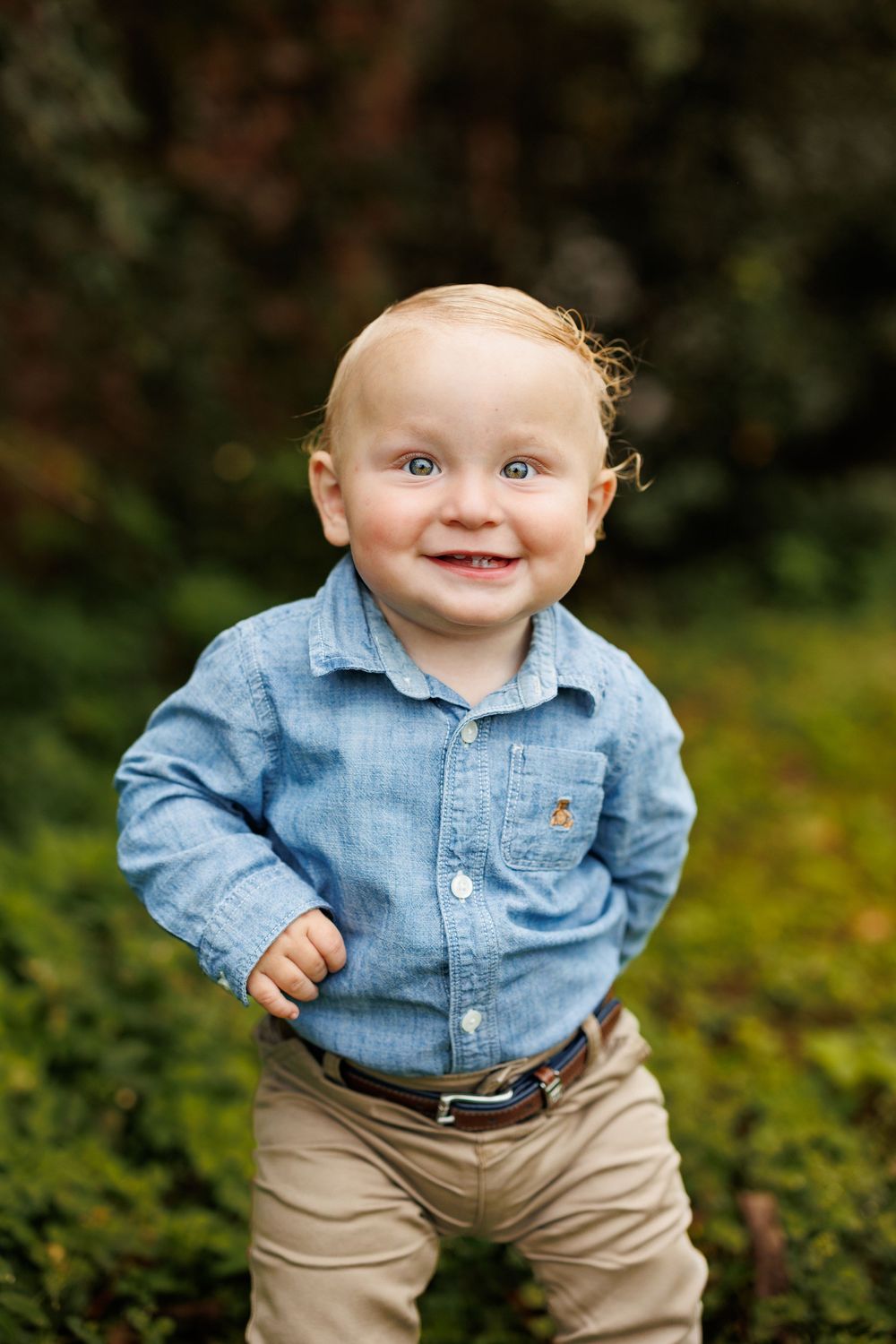 Baby in denim shirt and khaki pants smiles while standing in garden.