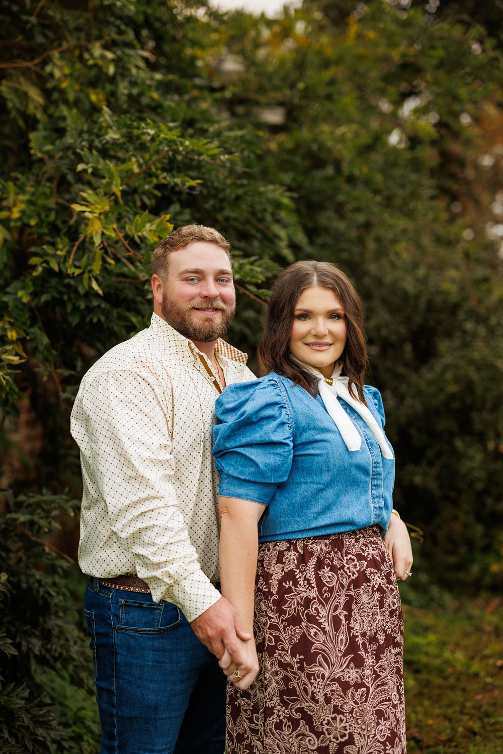 Couple in casual wear embraces while standing against green foliage background.