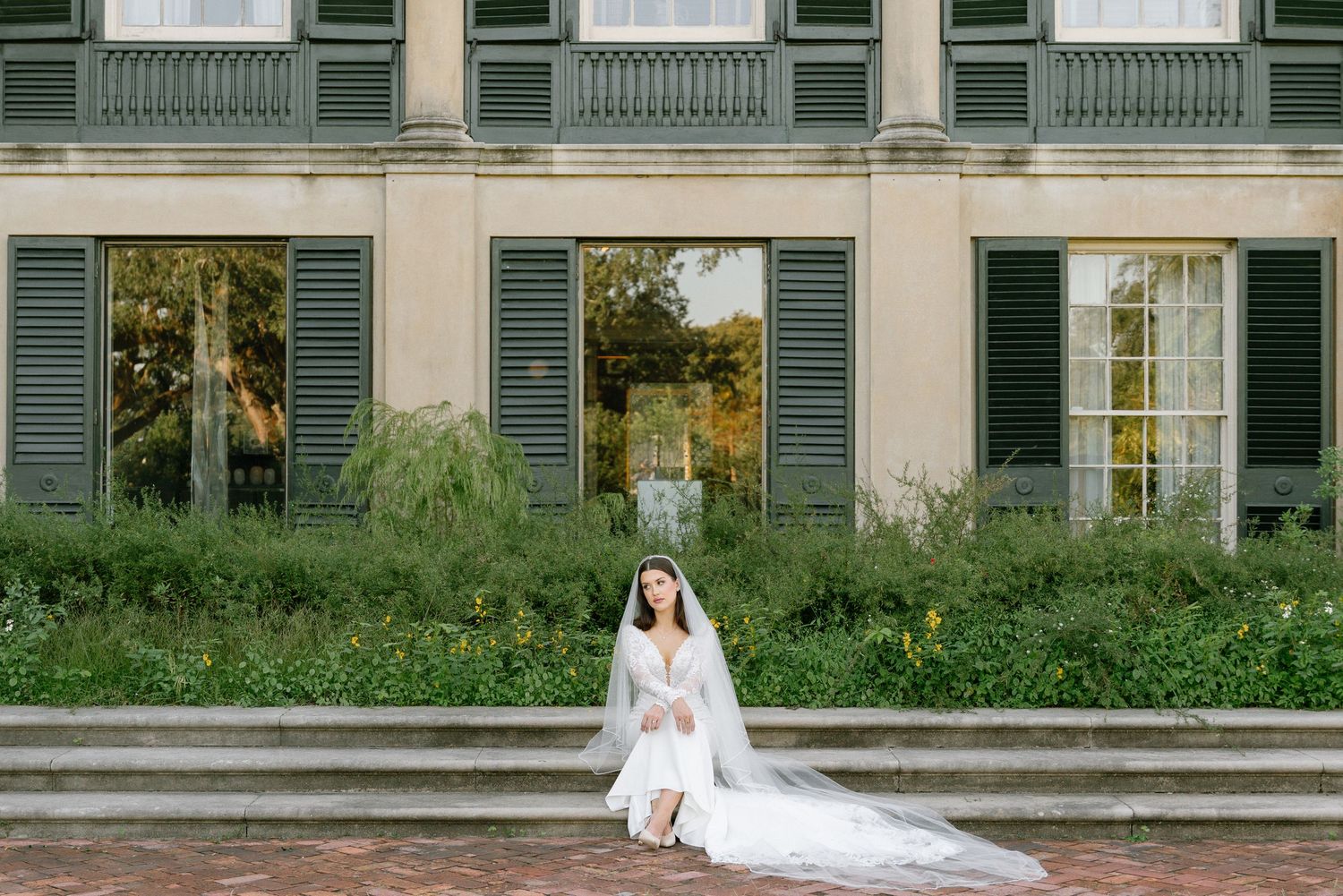 A bride in a white wedding dress poses in front of a historic building with green shuttered windows. New Orleans, longue vue gardens