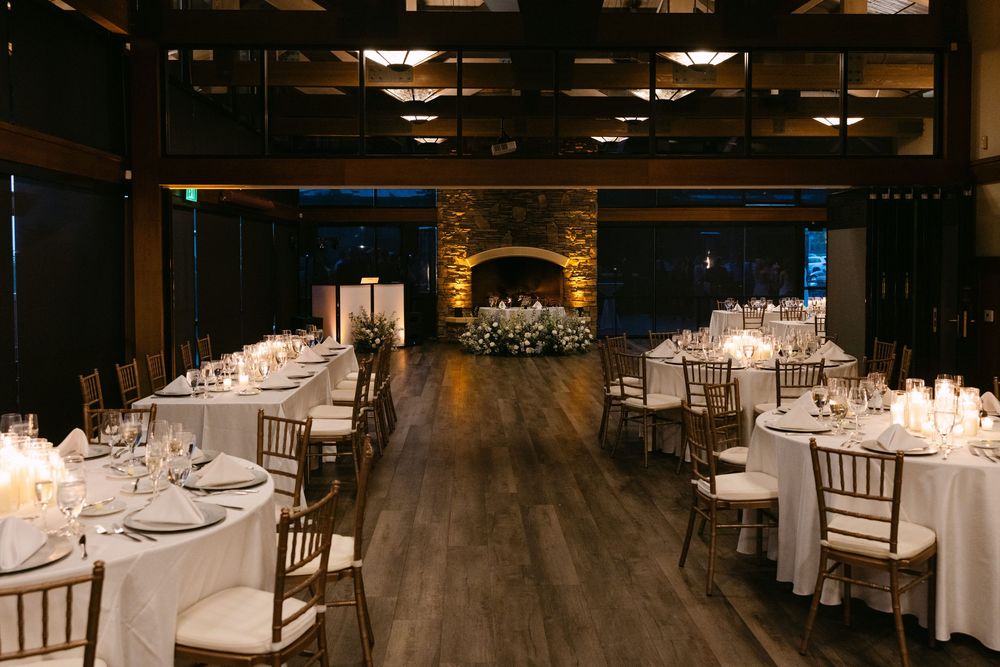 An elegant wedding reception venue with dark wood floors, stone fireplace, and white-clothed round tables set for dinner service.