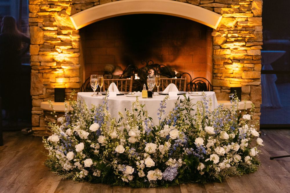 Elegant wedding reception table with stone fireplace backdrop and lush white floral arrangement display.