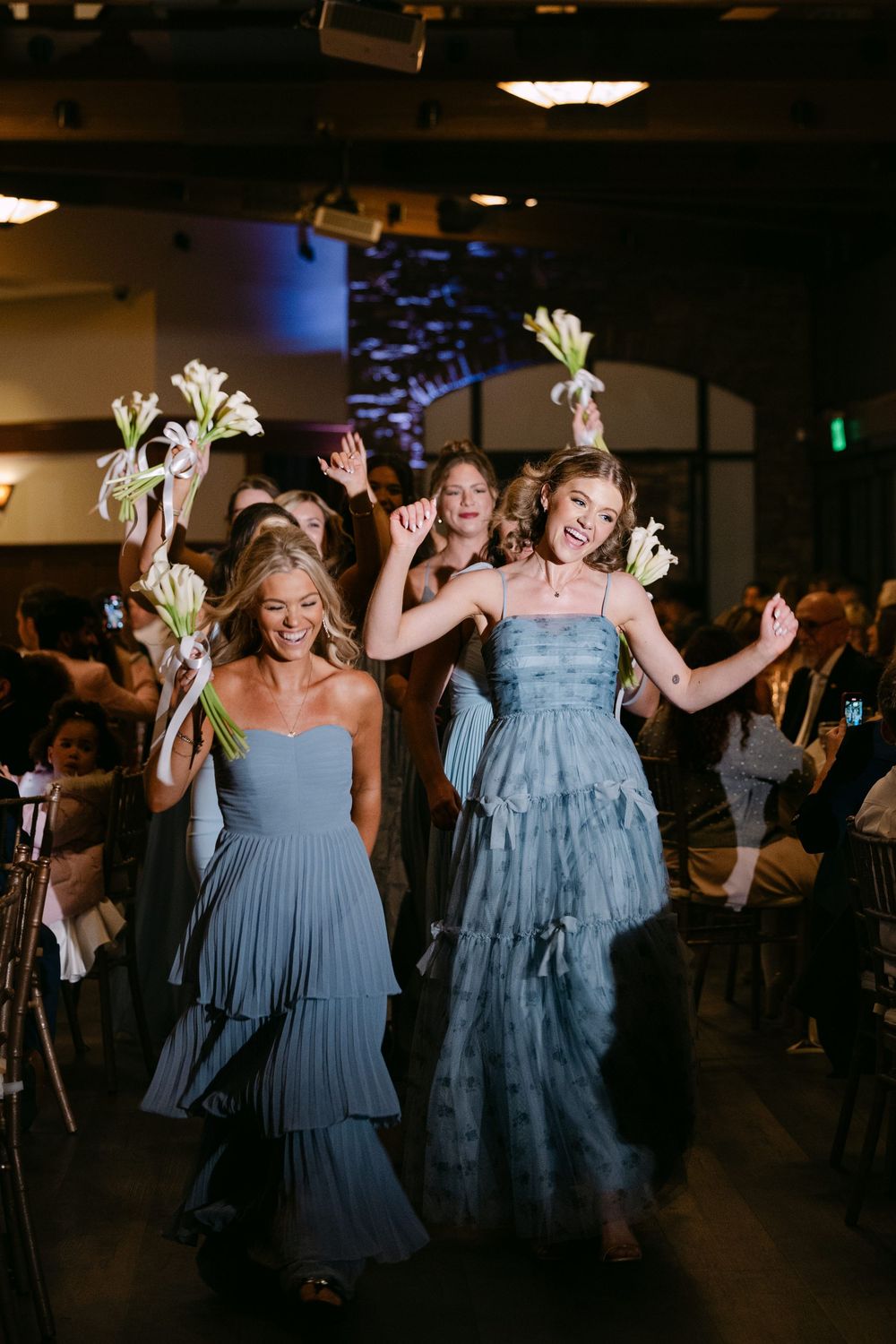 Wedding party members in blue dresses dance joyfully on the reception dance floor.