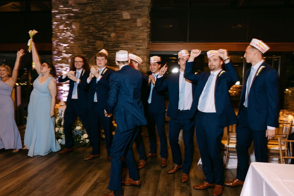 Wedding party and guests dancing together in formal attire at a rustic venue with stone walls and warm lighting.
