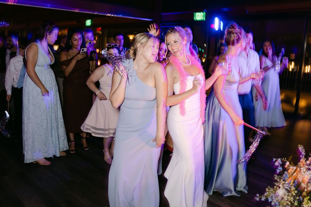Wedding party in white dresses dancing together on a dark dance floor illuminated by colorful lights.