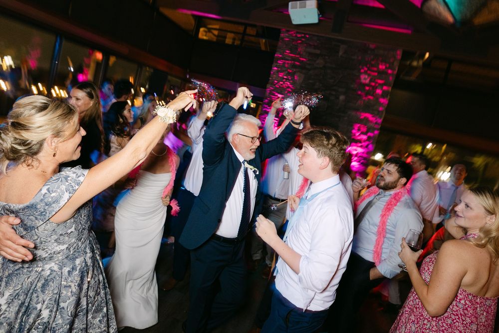 Wedding guests dance and celebrate on a crowded dance floor under pink and purple lighting at an evening reception.