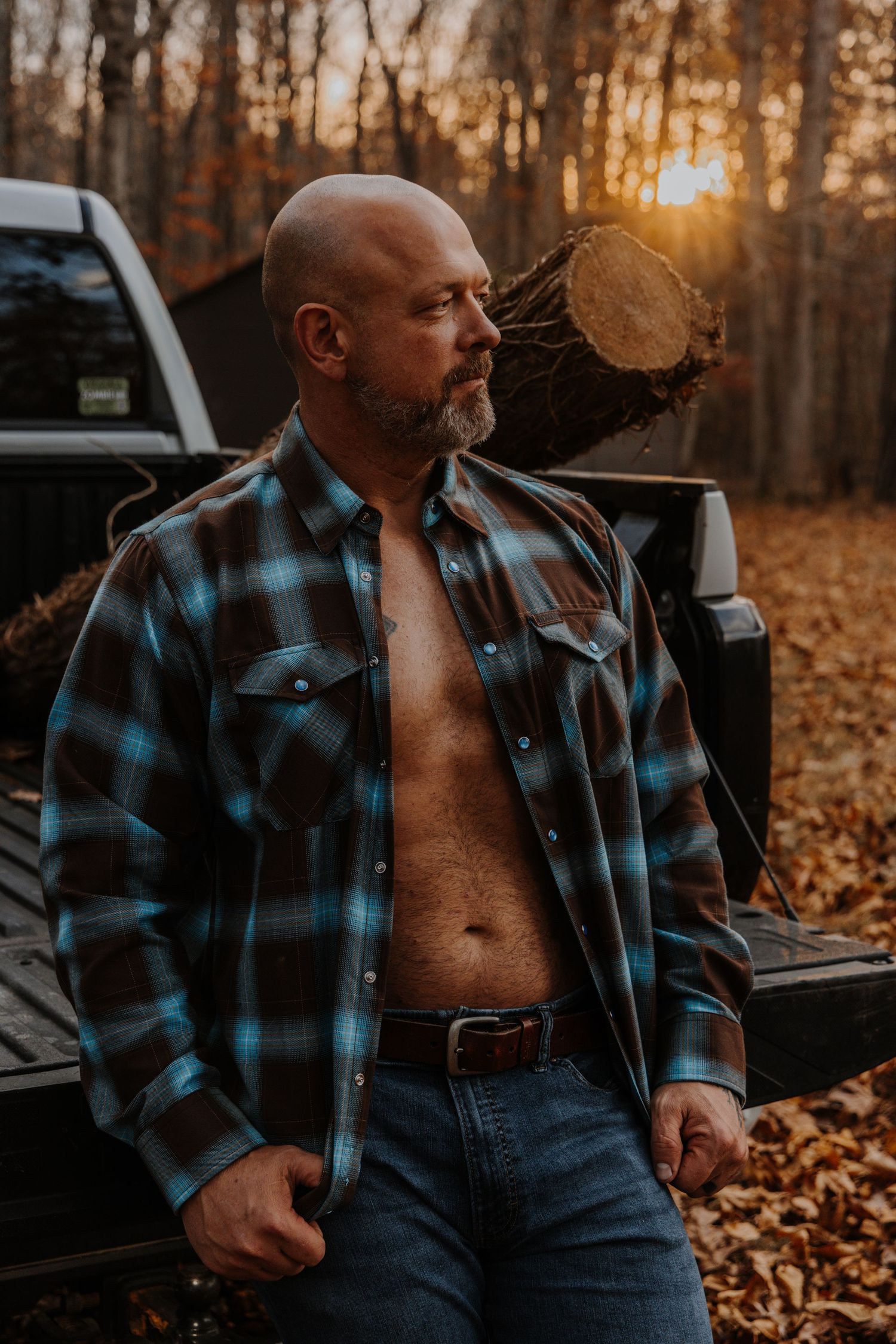 Person in plaid flannel shirt and jeans sits on fallen leaves during golden sunset in autumn forest.