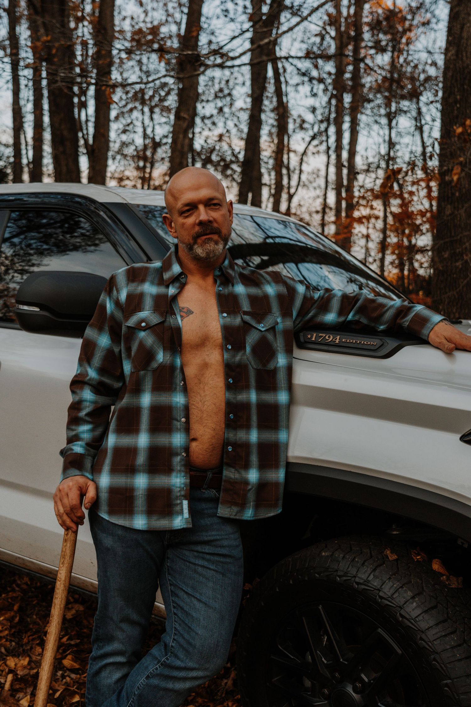 Person wearing plaid flannel shirt and jeans stands beside white vehicle in autumn forest setting.