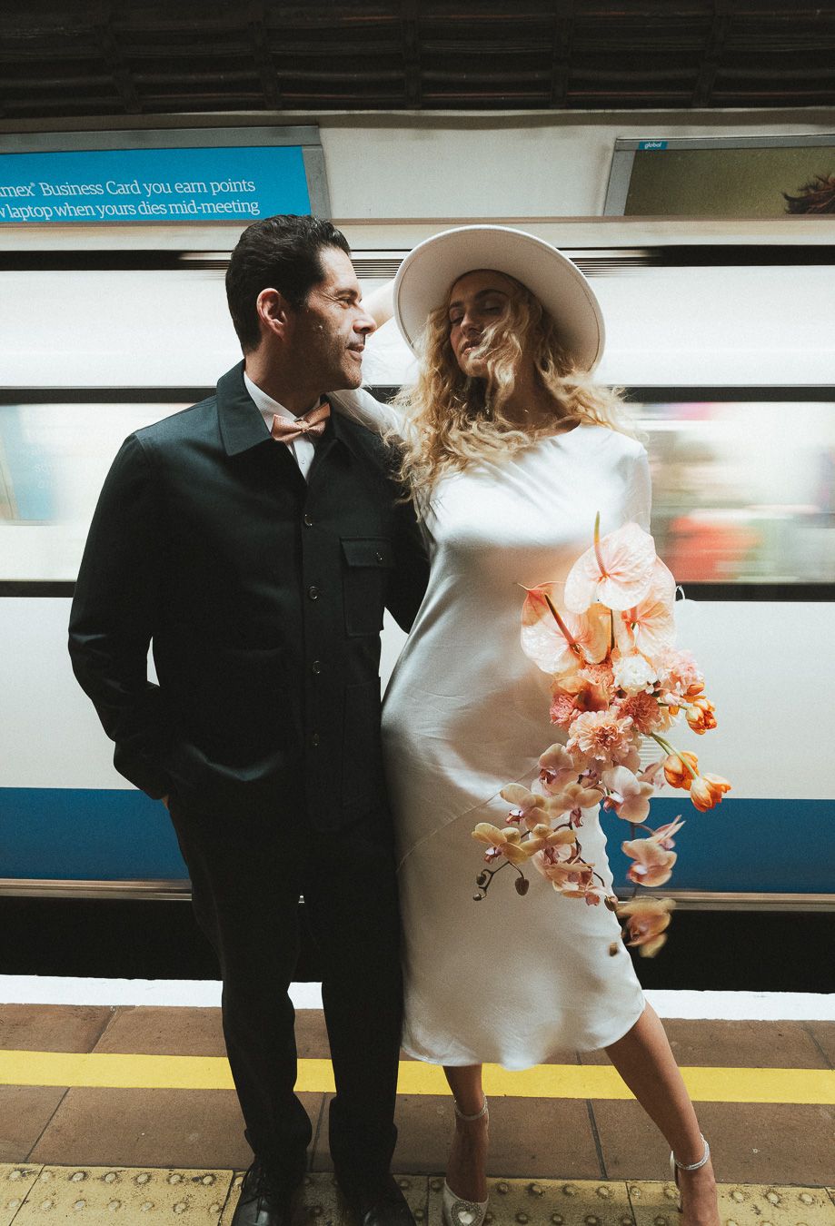 A couple stands on a  London subway platform in formal attire with peach-coloured flowers creating a romantic moment.