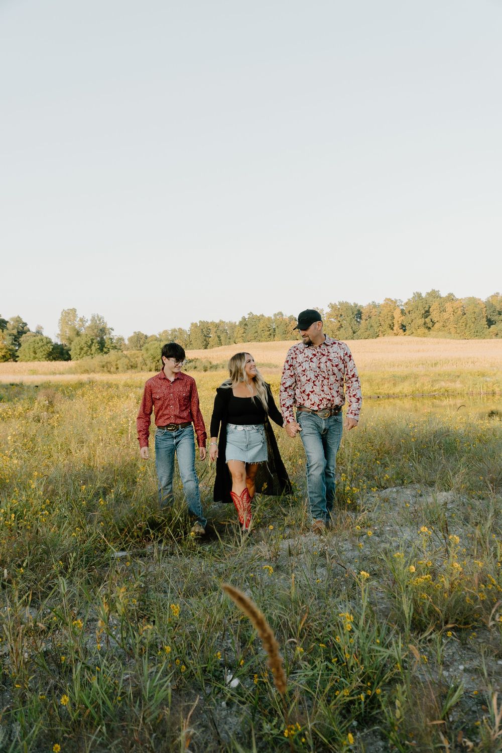Three people walk together through a golden field at sunset with tall grass and wildflowers in the foreground.