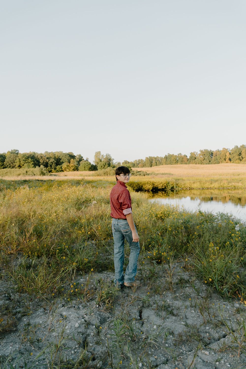 Person in red sweater and jeans stands on sandy bank near calm pond and autumn trees at golden hour.