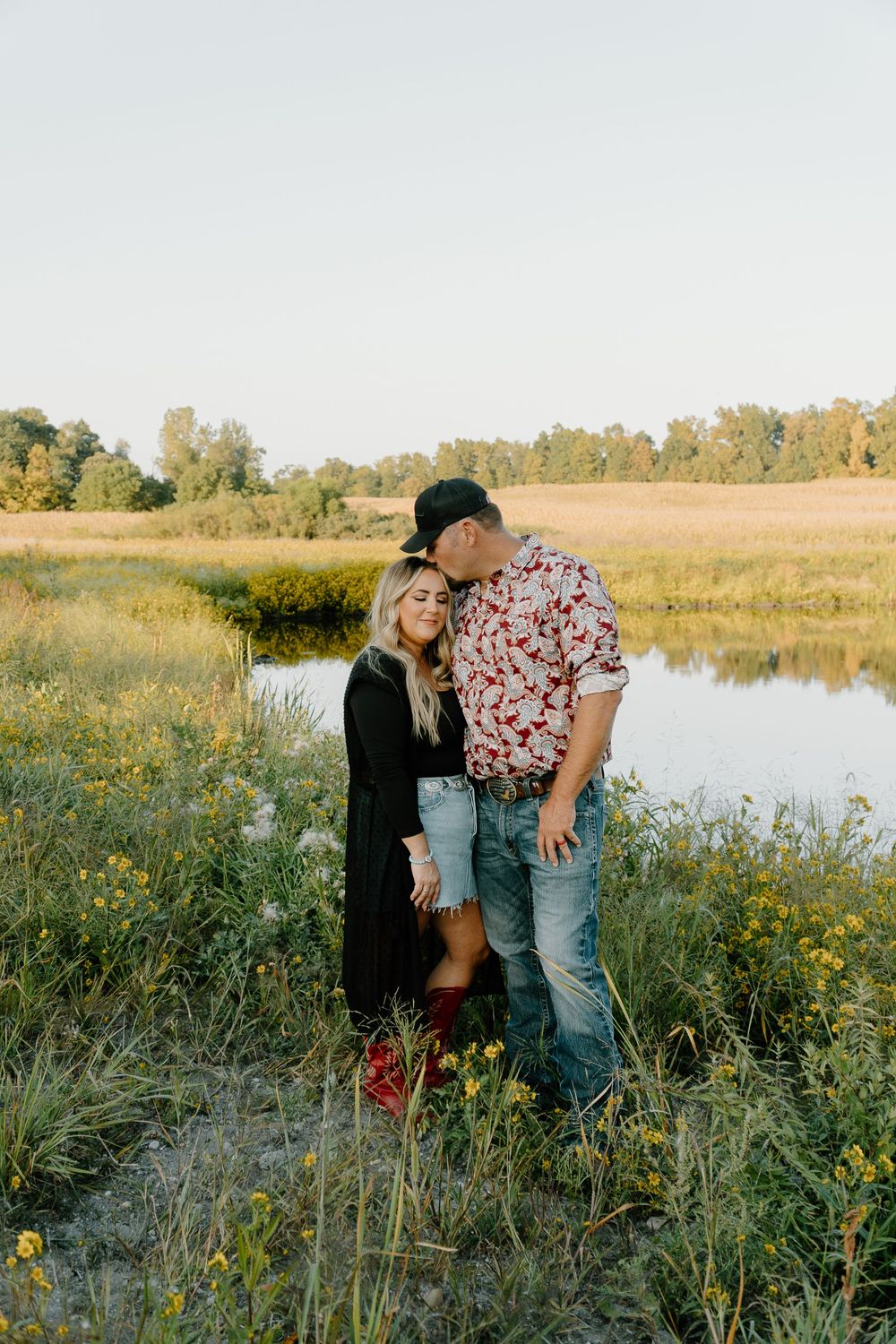 A couple shares intimate moments in a golden field near a pond during sunset.