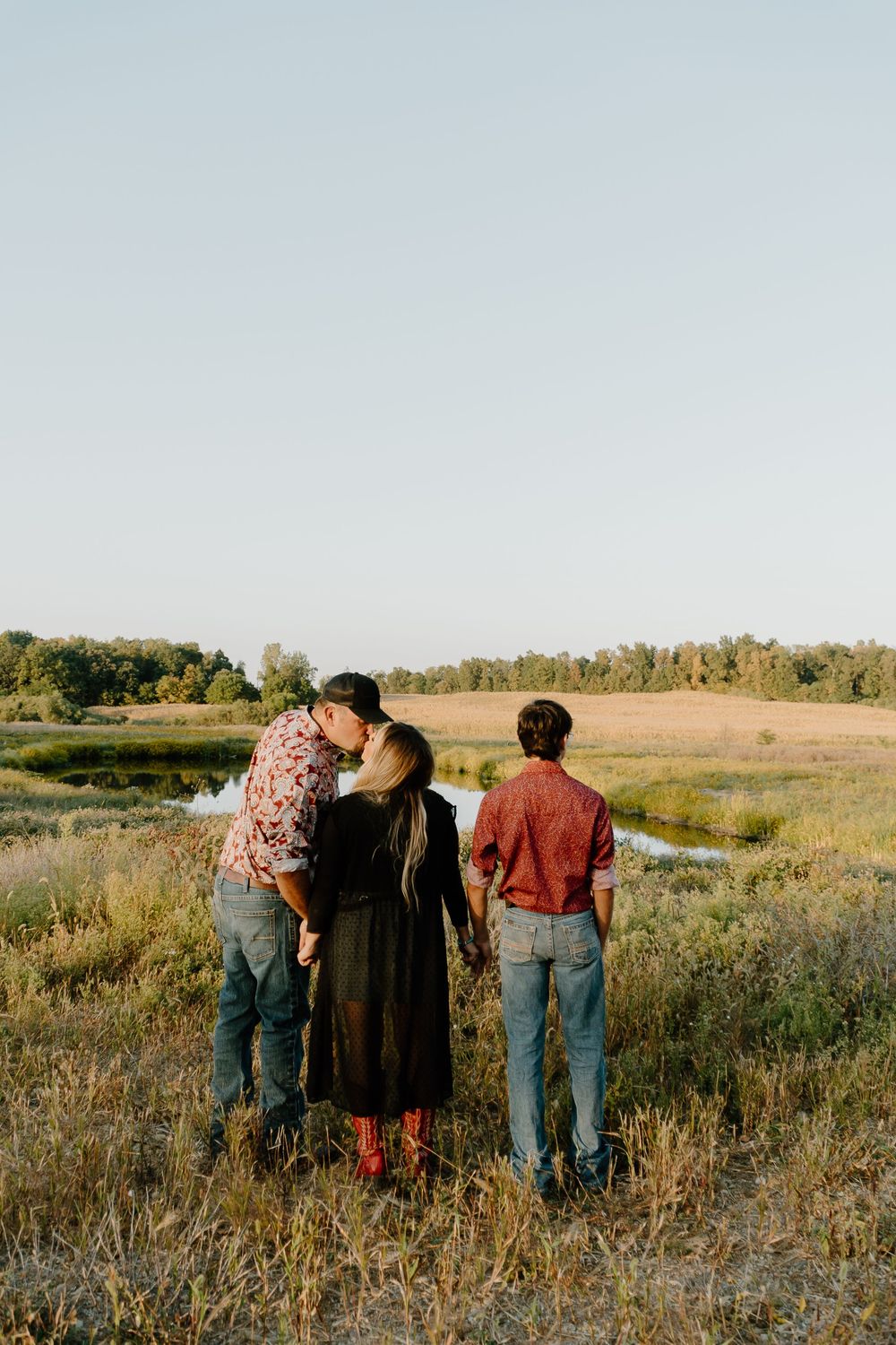 Three people stand together in a golden field at sunset with trees visible in the background.