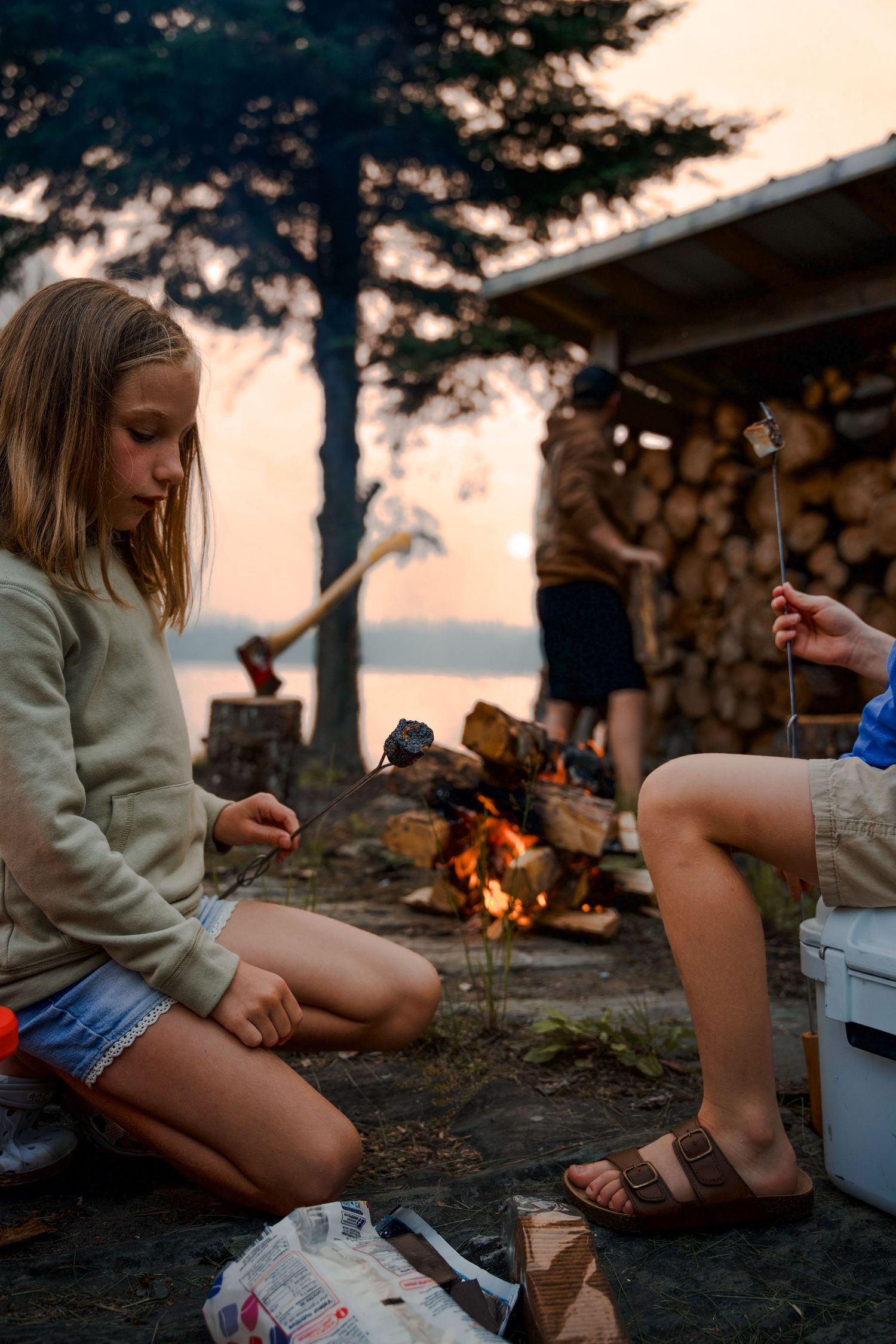 Cozy campfire scene with people relaxing at sunset near stacked firewood.