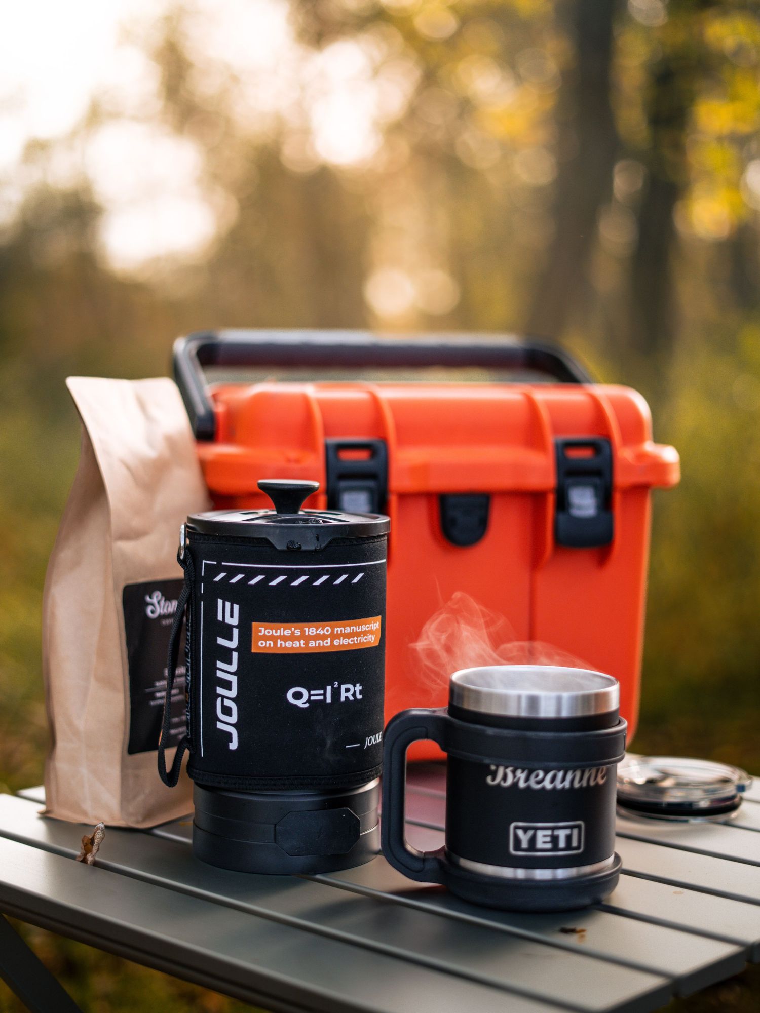 Outdoor coffee setup with red cooler and black Yeti mugs on portable table.