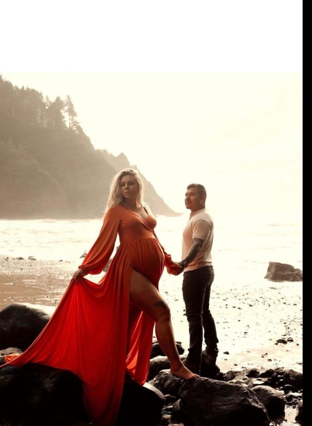 A couple in a romantic pose on a misty beach, with the woman wearing a flowing red dress against the rocky coastline.