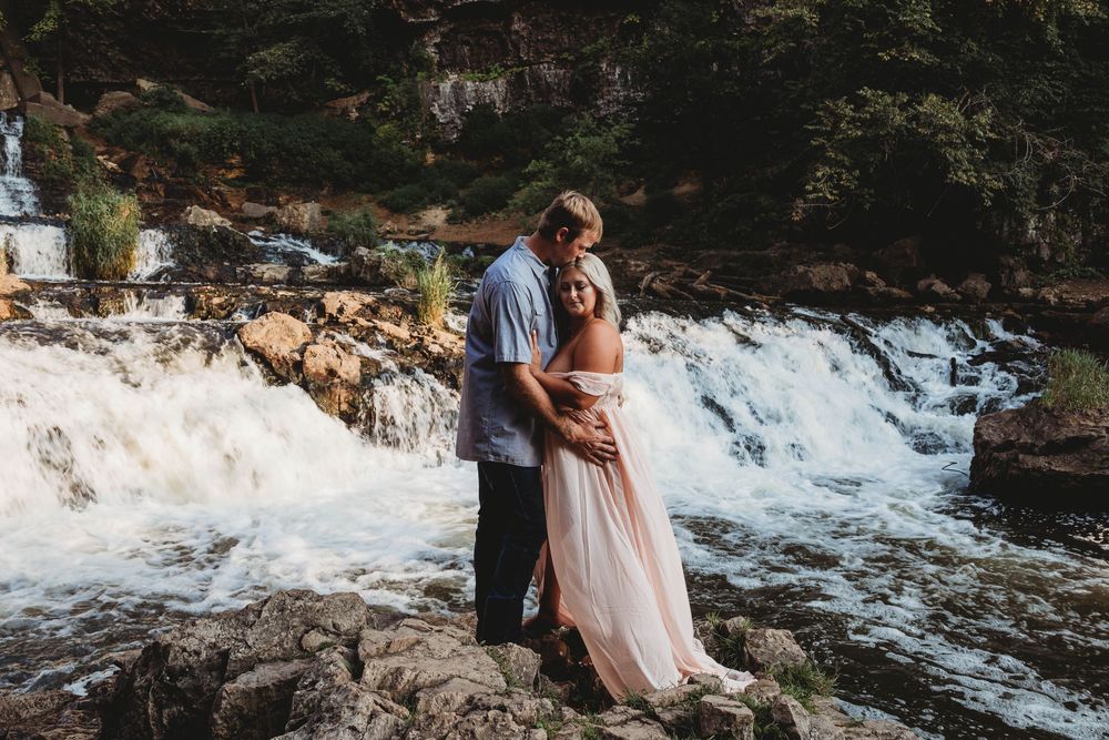 Couple embracing romantically on rocks in front of a cascading waterfall, woman in flowing pink dress.