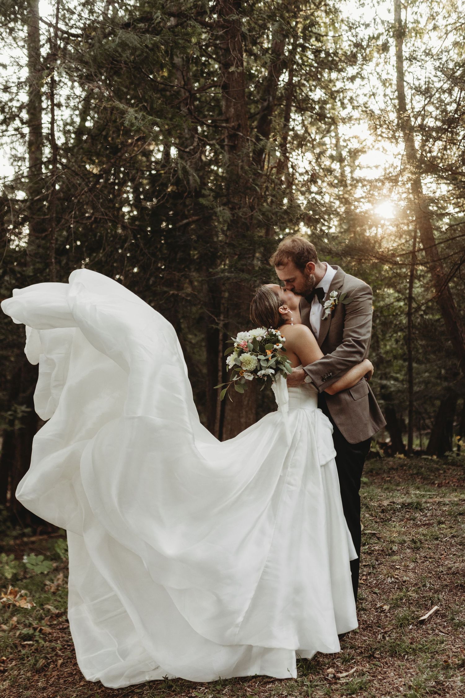 A couple in wedding attire walks through a sunlit forest holding hands as the bride's dress flows behind them.