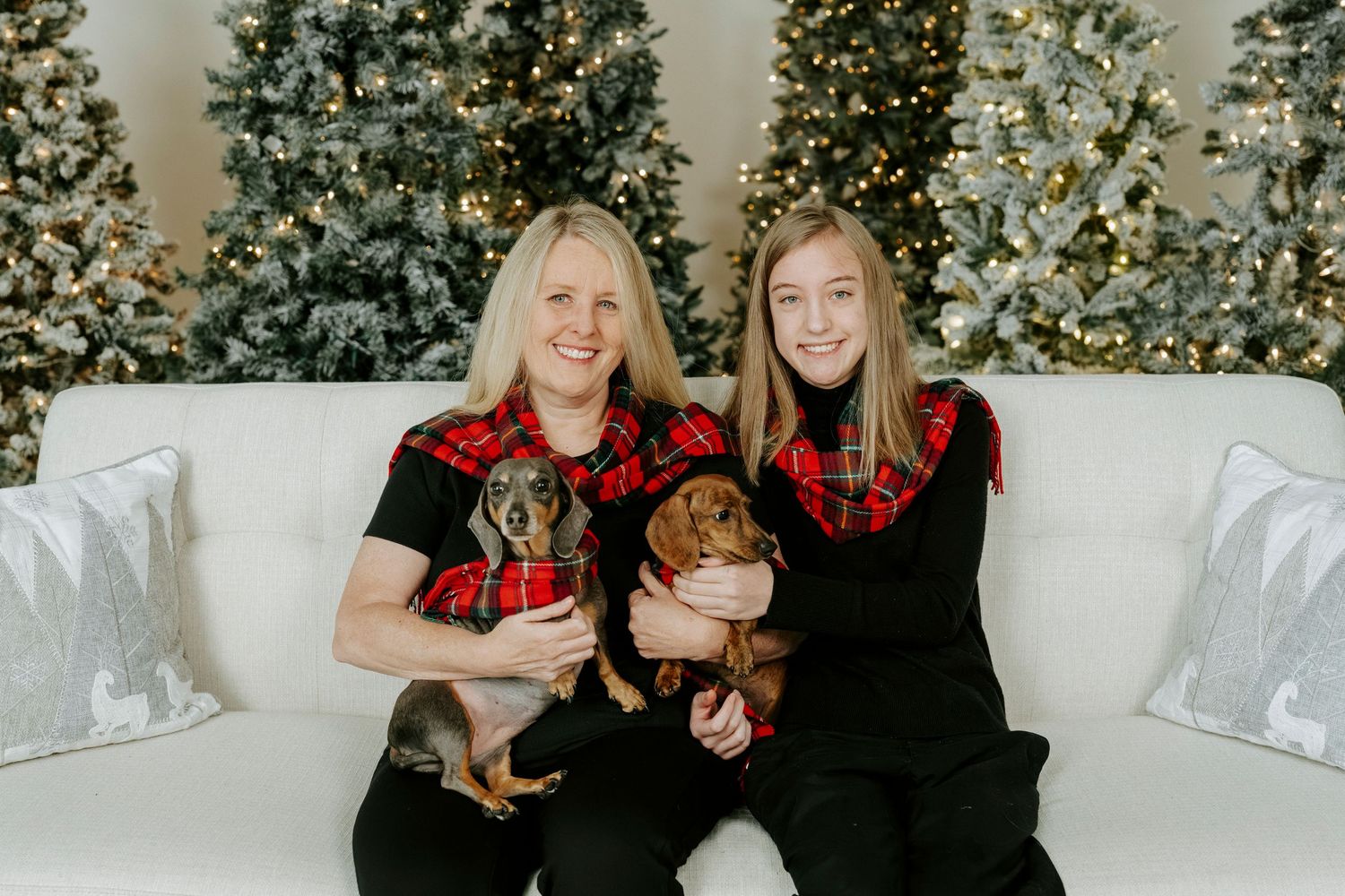 Two people wearing black and red outfits pose together on a white couch with decorated Christmas trees in background.