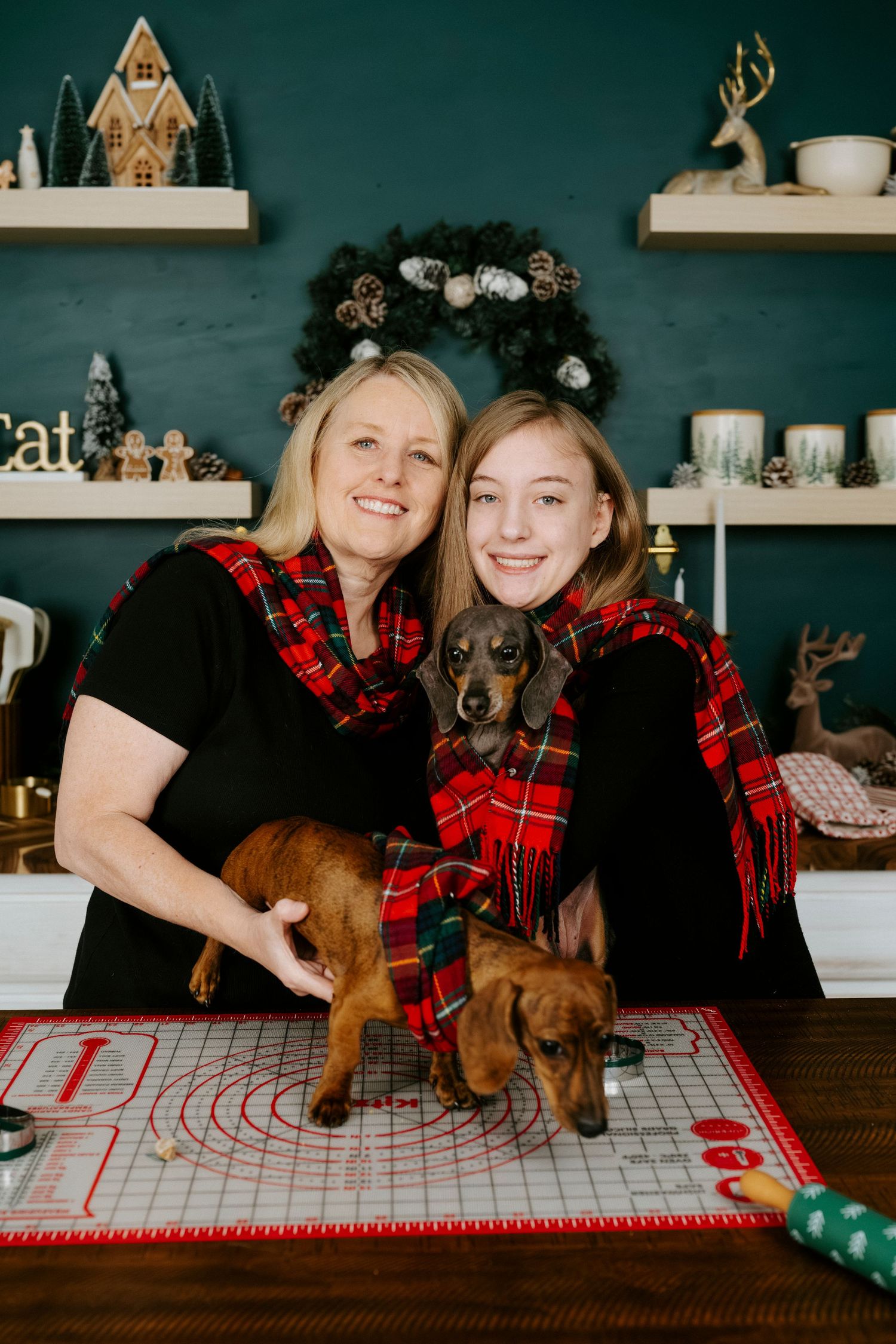 Two dachshund dogs pose with their owners in matching red plaid holiday outfits against a green wall with festive decor.
