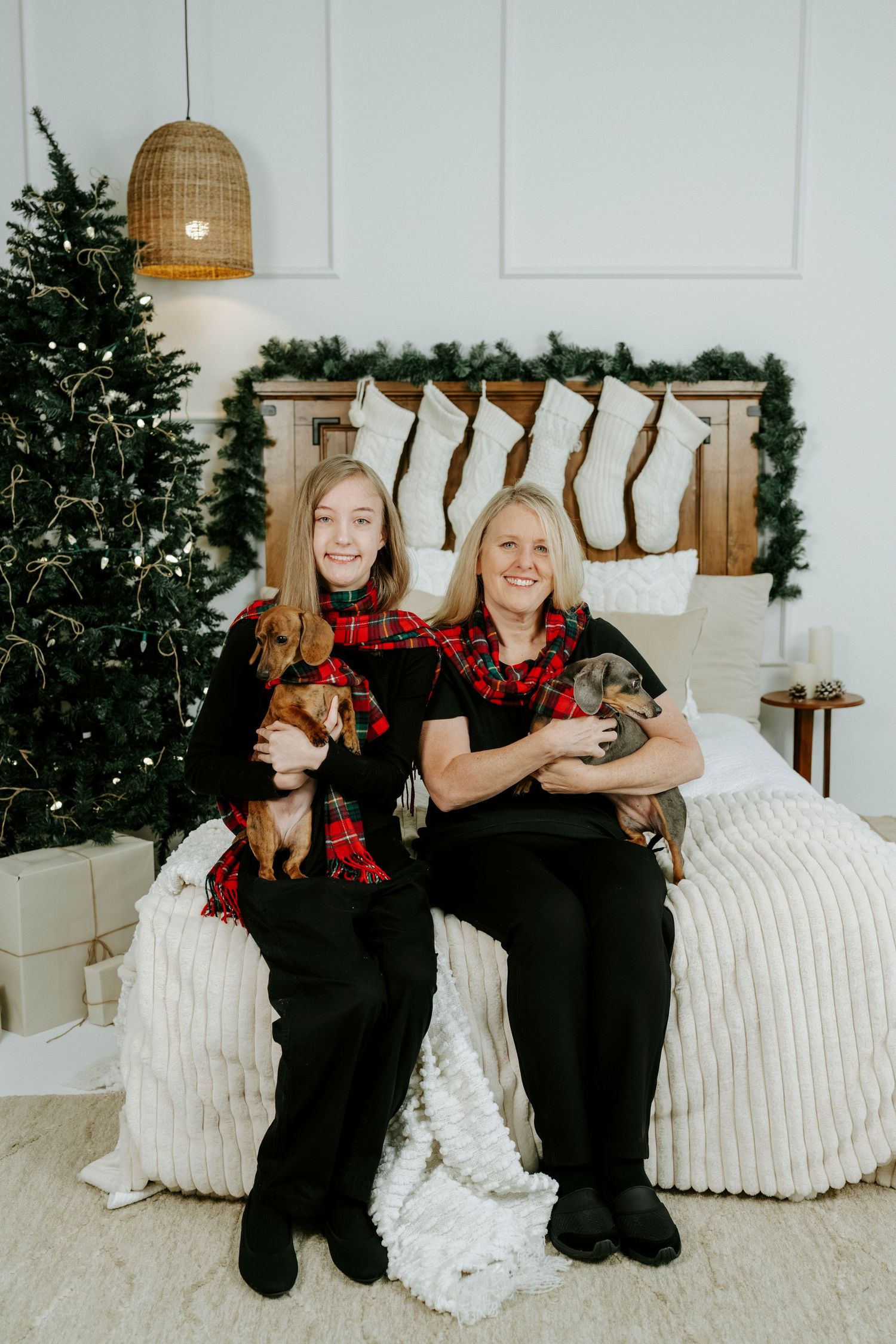 Two people in matching black outfits sit on a bed with small dogs against a Christmas backdrop with stockings.